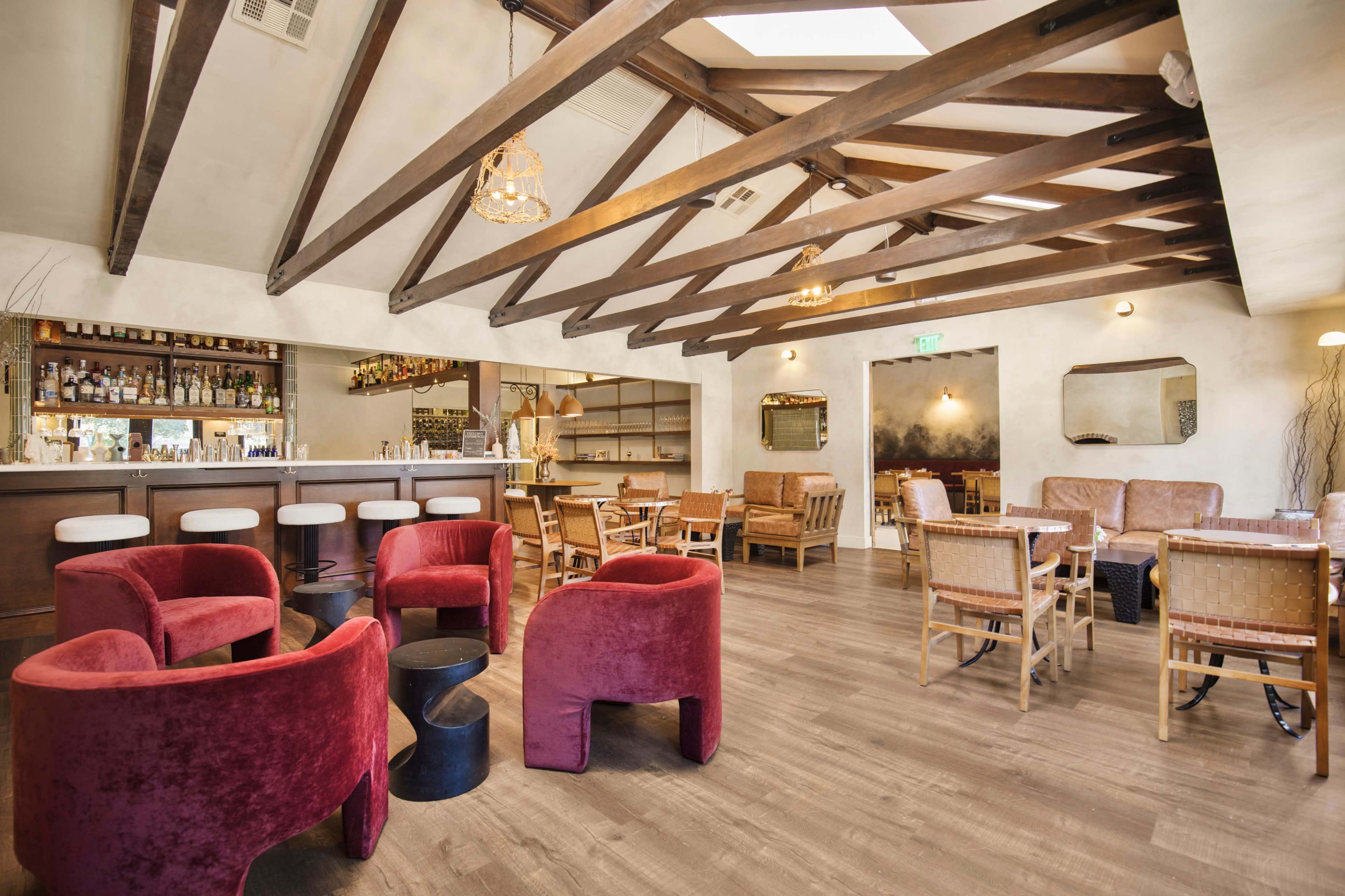 The image shows a modern dining space featuring a bar, seating areas with red chairs, wooden beams, and a warm ambiance.