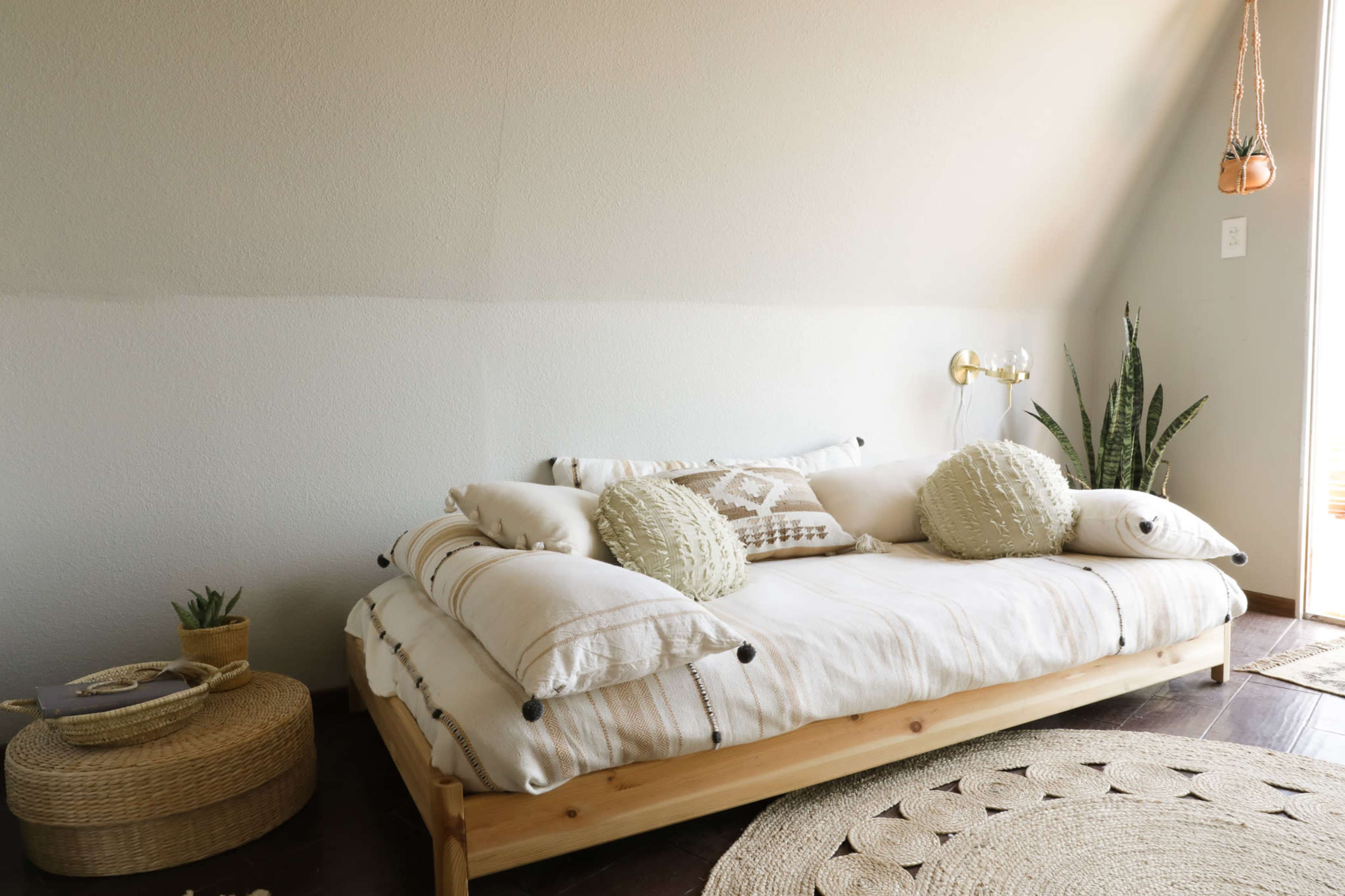 A minimalist bedroom with a wooden daybed adorned with neutral-colored cushions and a round woven rug beside it.