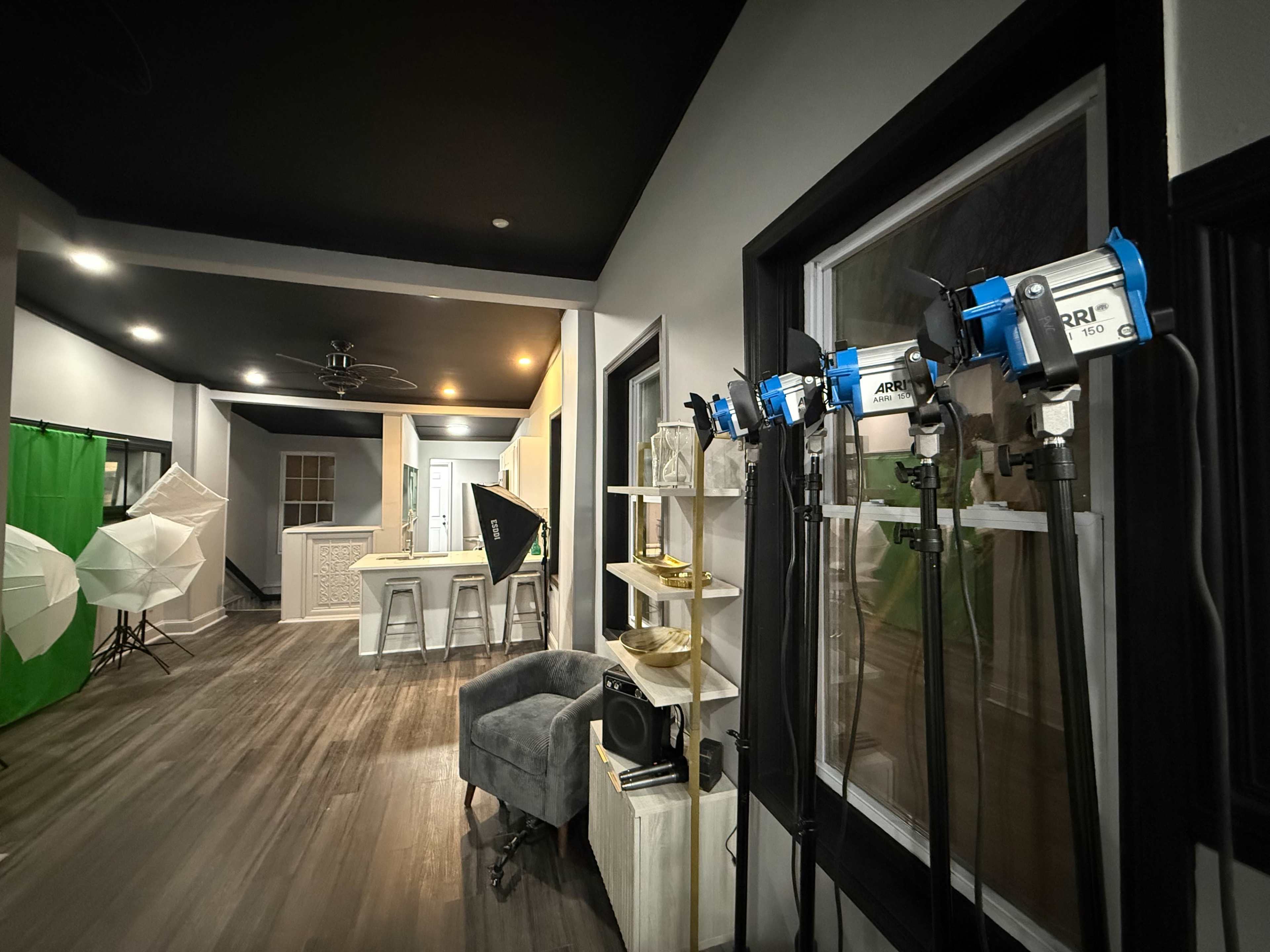 The image shows a well-lit photography studio with various lighting equipment on stands, a seating area, and a backdrop setup in the background.