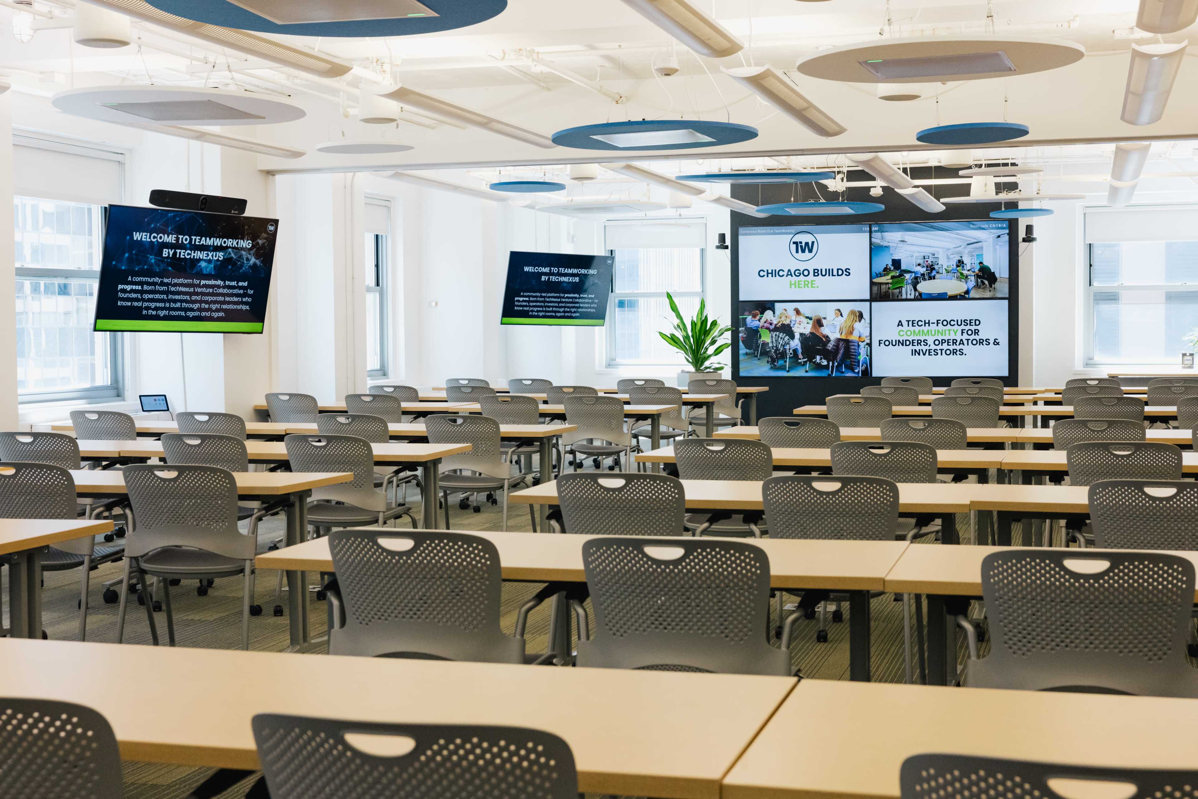 The image shows a bright, modern classroom with rows of tables and chairs, featuring screens displaying the text "Welcome to Transforming Tech" and "Chicago Builds."
