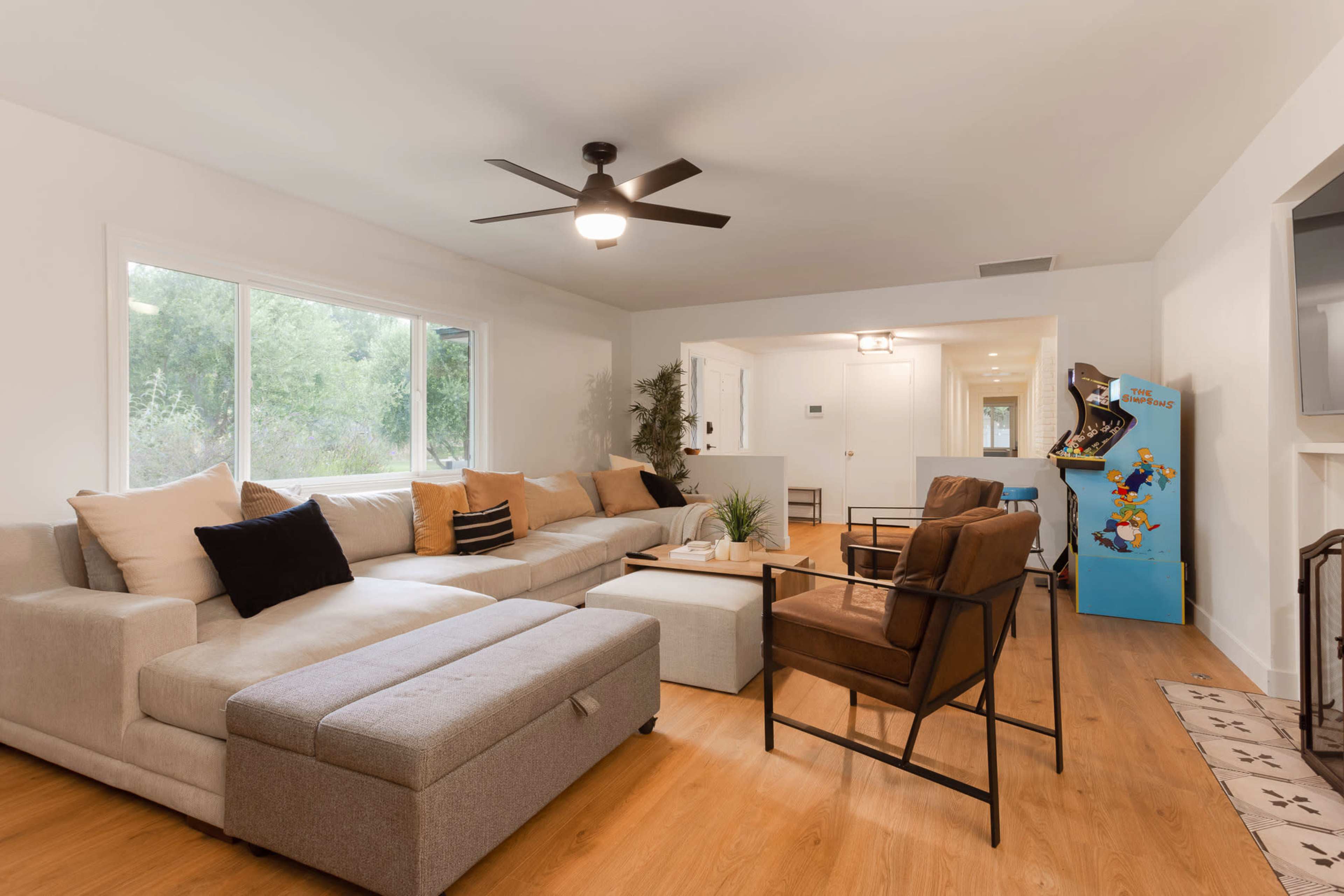 The living room features a large beige sectional sofa, two brown armchairs, a coffee table, and a colorful arcade machine in the corner, with natural light streaming through a large window.