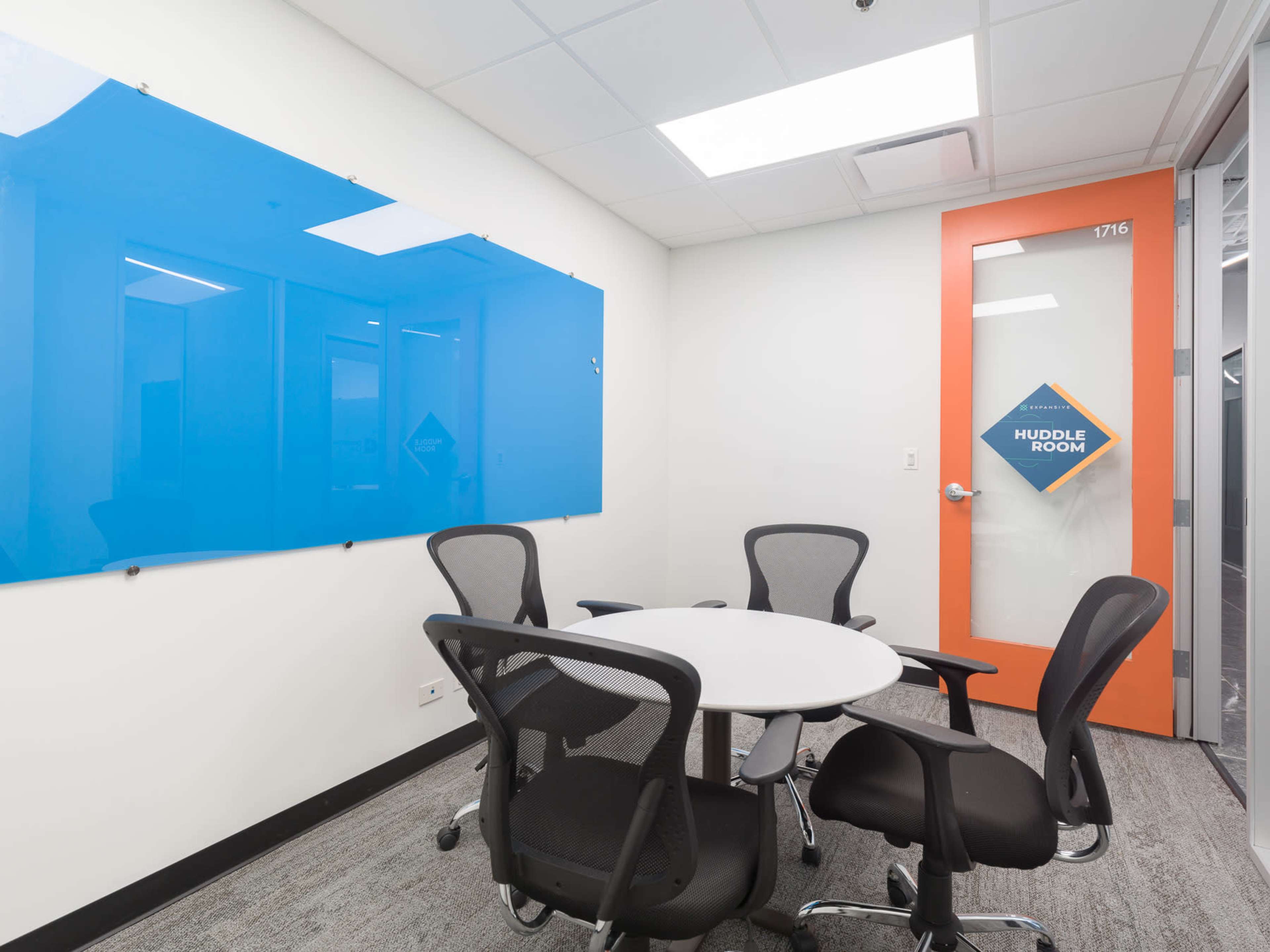 The image shows a small meeting room with a round table, four black chairs, a blue whiteboard on the wall, and an orange door labeled "Huddle Room."