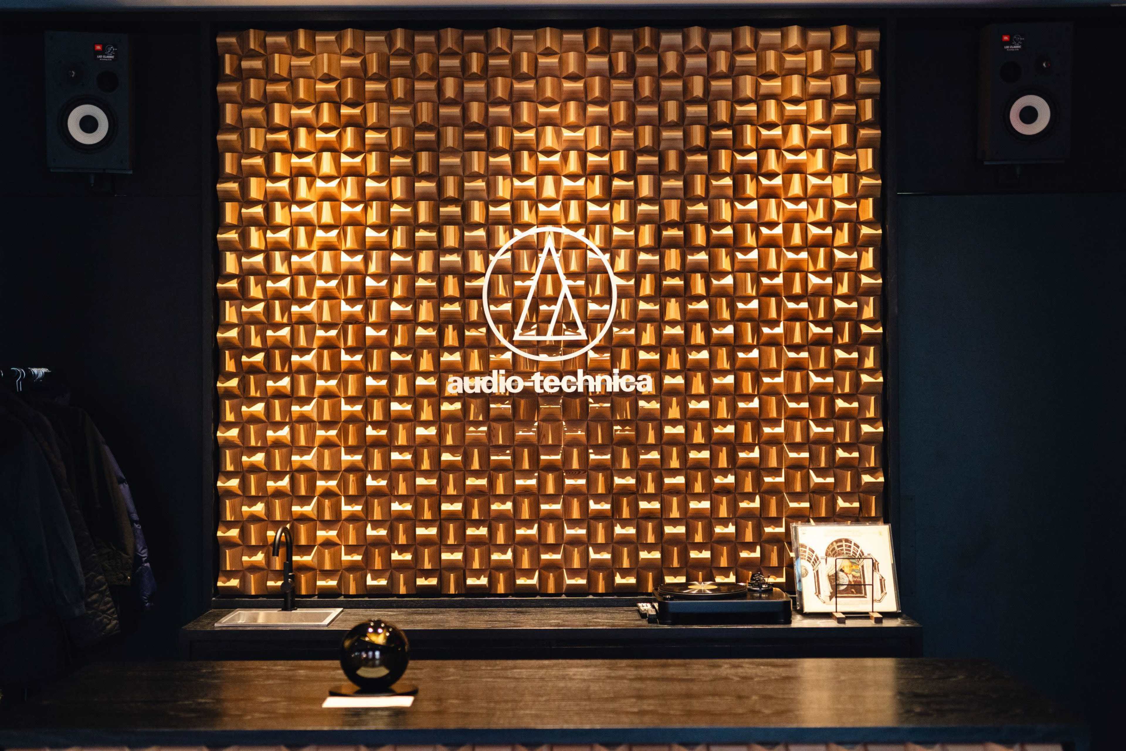 The image features a modern interior with a wooden wall displaying the audio-technica logo, flanked by two speakers, and a dark countertop with a record player and a framed photo.