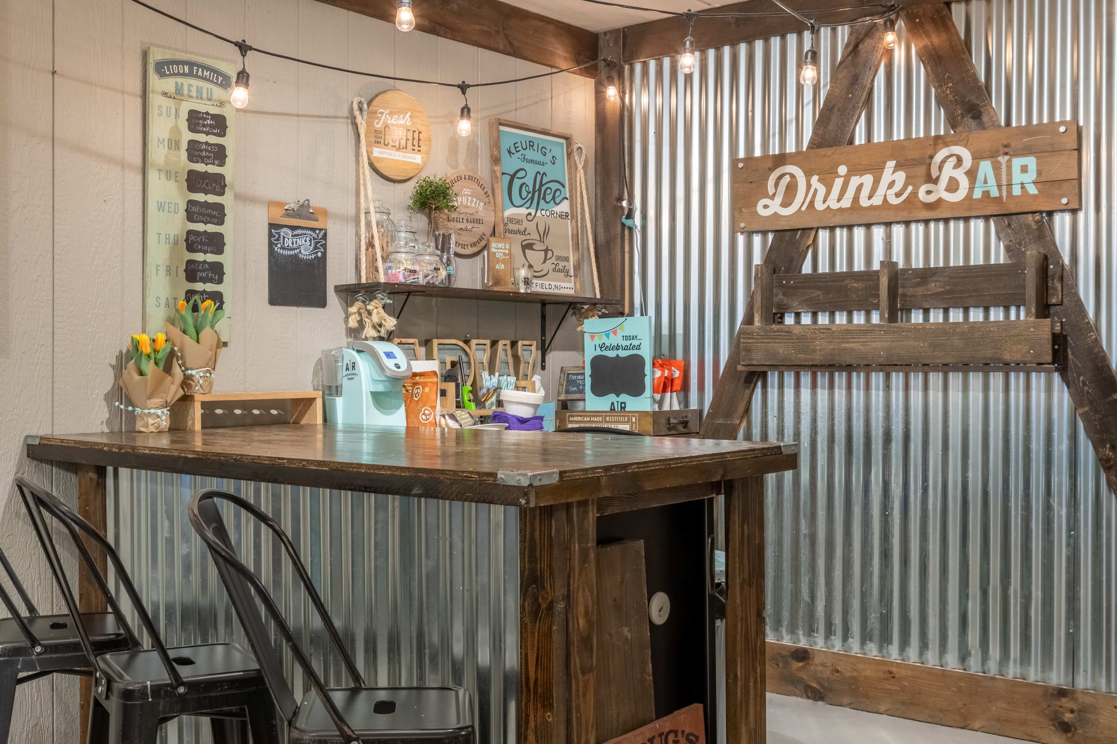 A rustic drink bar with a wooden counter, metal wall paneling, decorative signs, and string lights overhead.