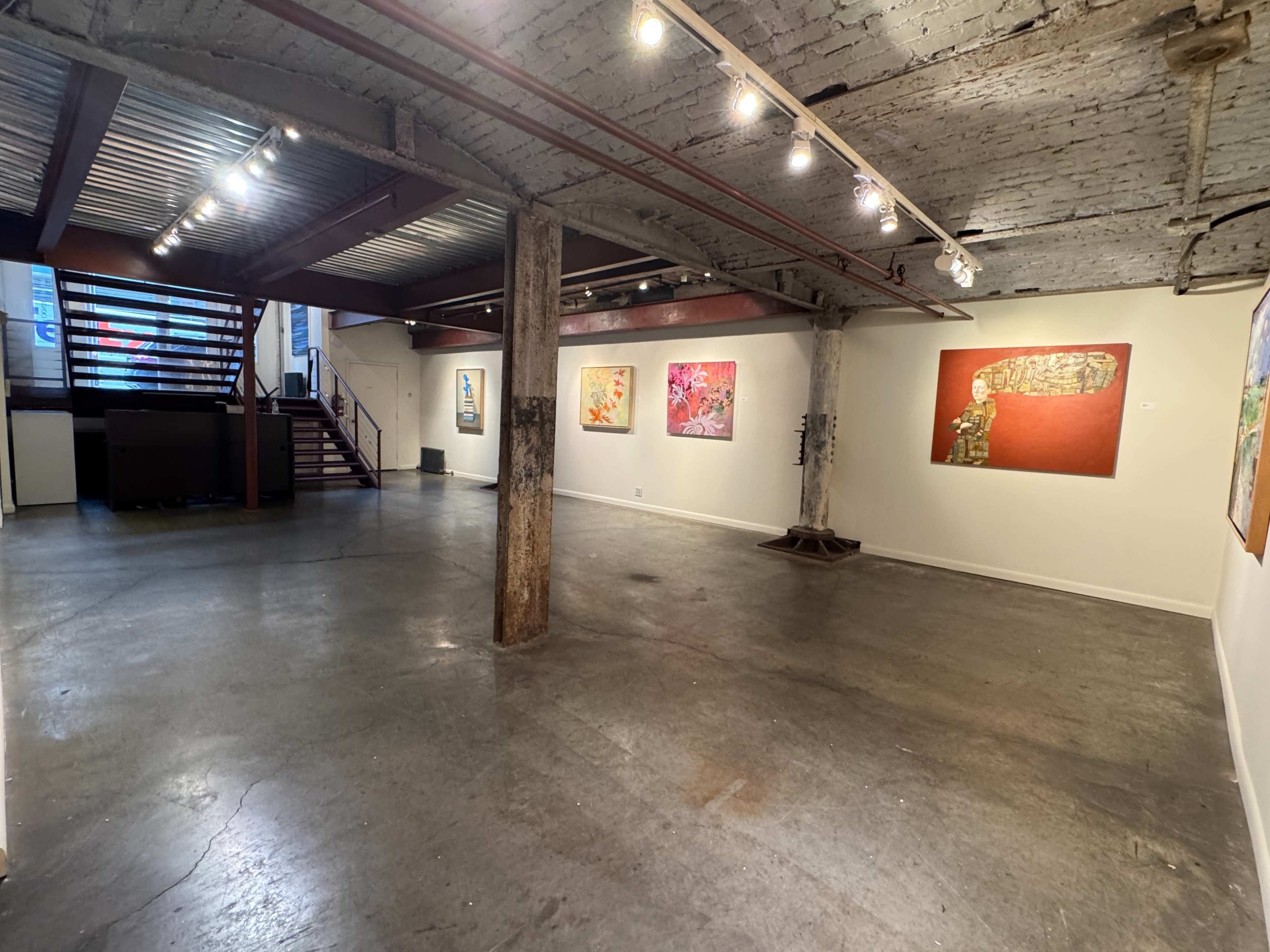 The image shows an art gallery with several paintings displayed on white walls, exposed brick and wooden beams, and a staircase in the background.