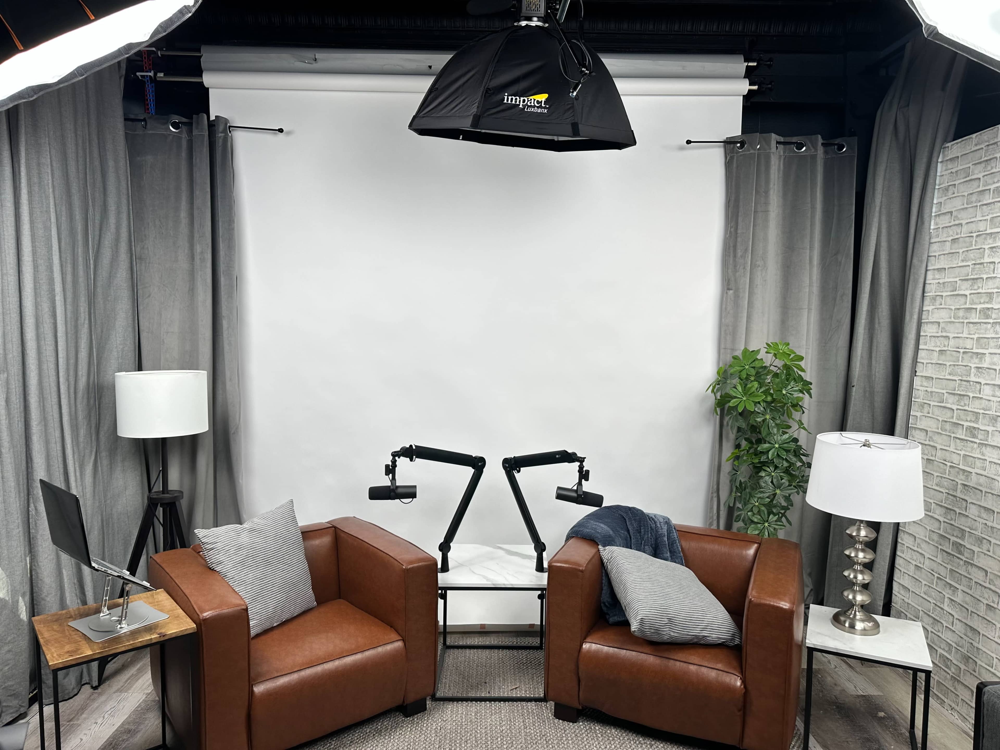 A studio setup featuring two brown armchairs, a small table with a laptop, and various lighting equipment against a white backdrop.
