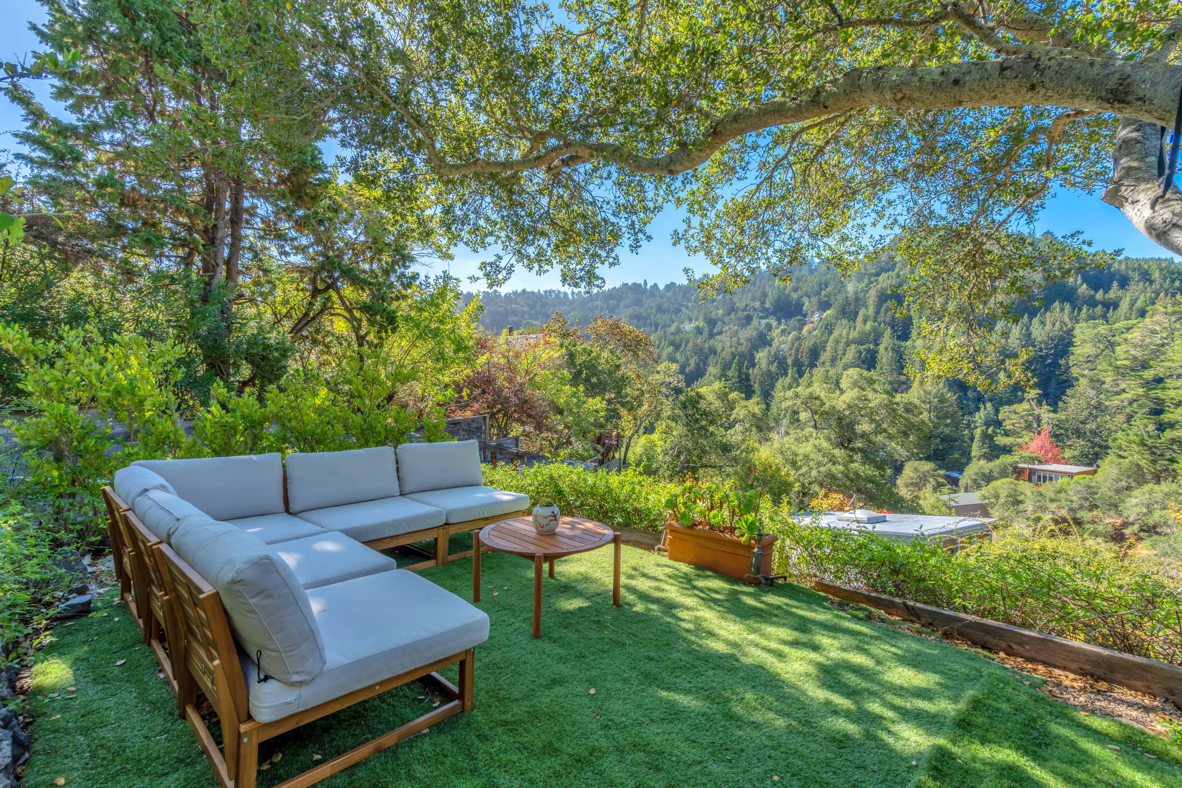 A comfortable outdoor seating area with a sofa and table is situated on a green lawn surrounded by trees and a scenic view of rolling hills.