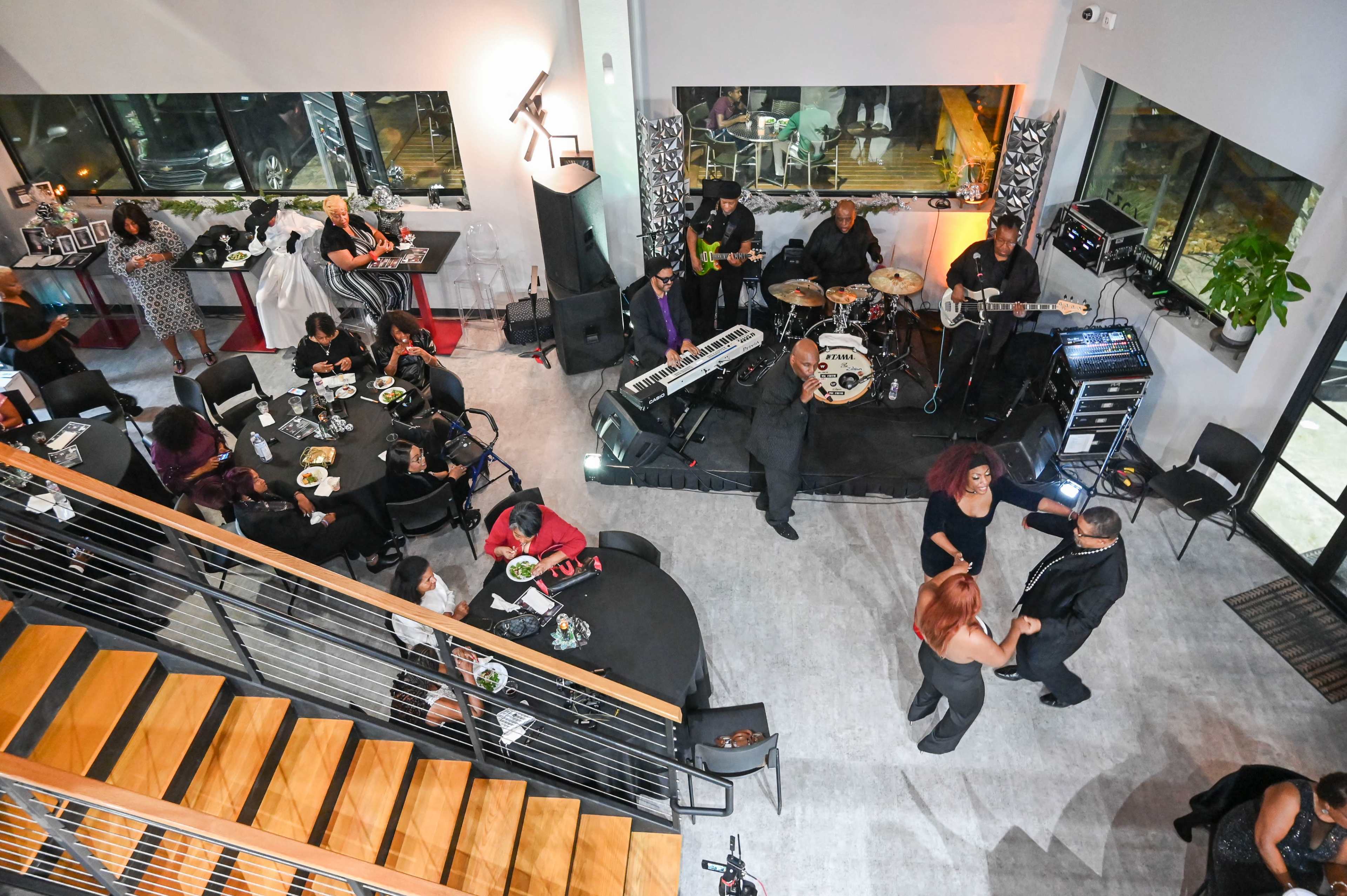 A lively indoor event features a band playing music while guests enjoy food and dancing at tables and on a dance floor.