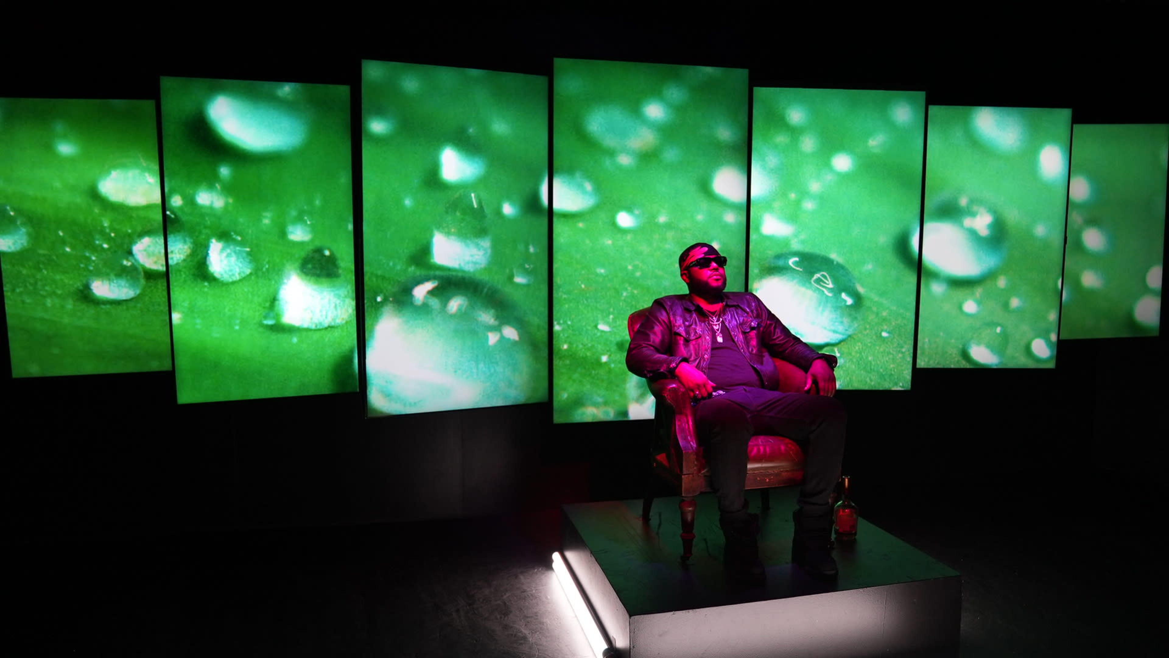 A man sits confidently in a chair against a backdrop of large screens displaying green leaves with water droplets.