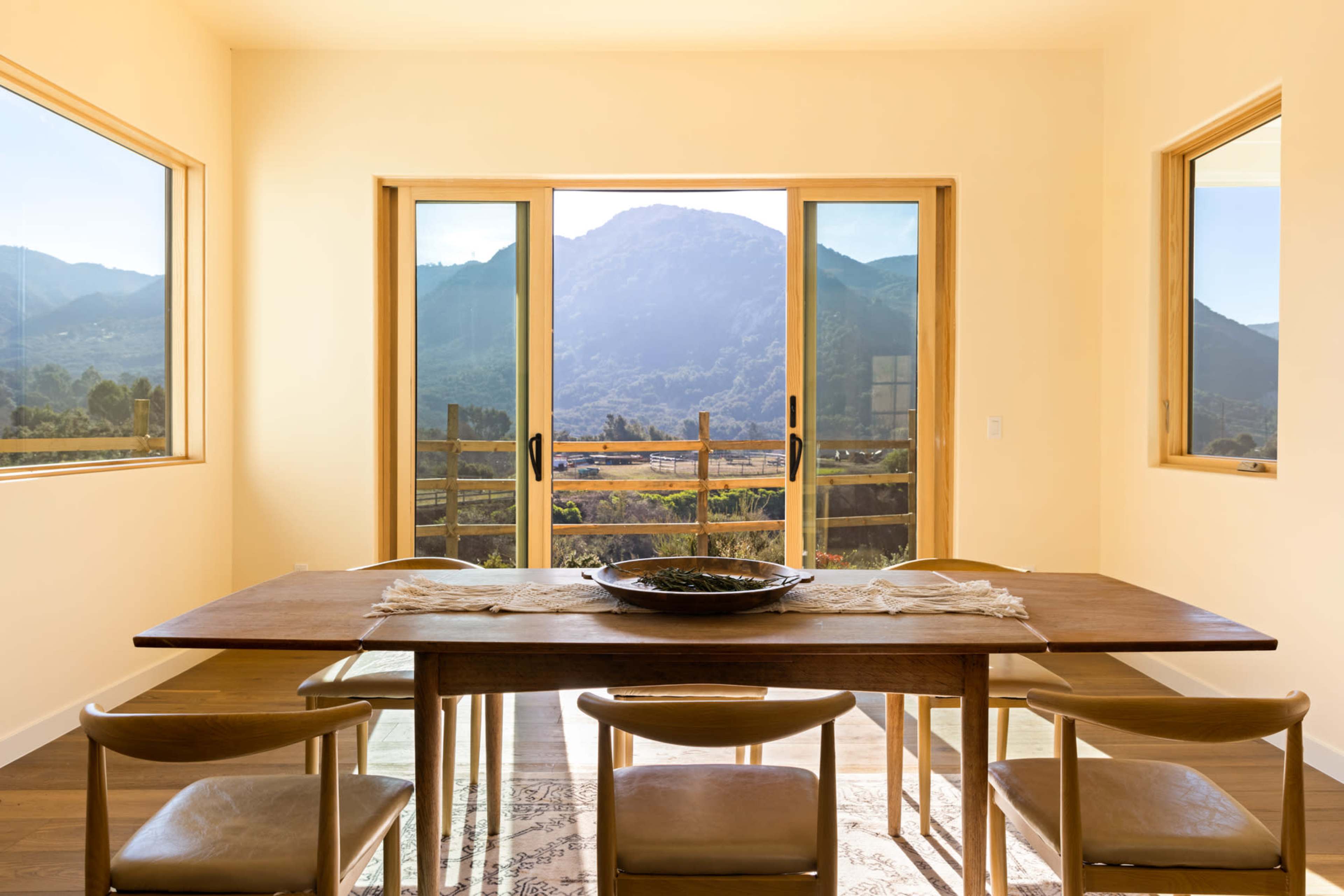 A wooden dining table with chairs is positioned in a brightly lit room that overlooks a mountain view through large glass doors.