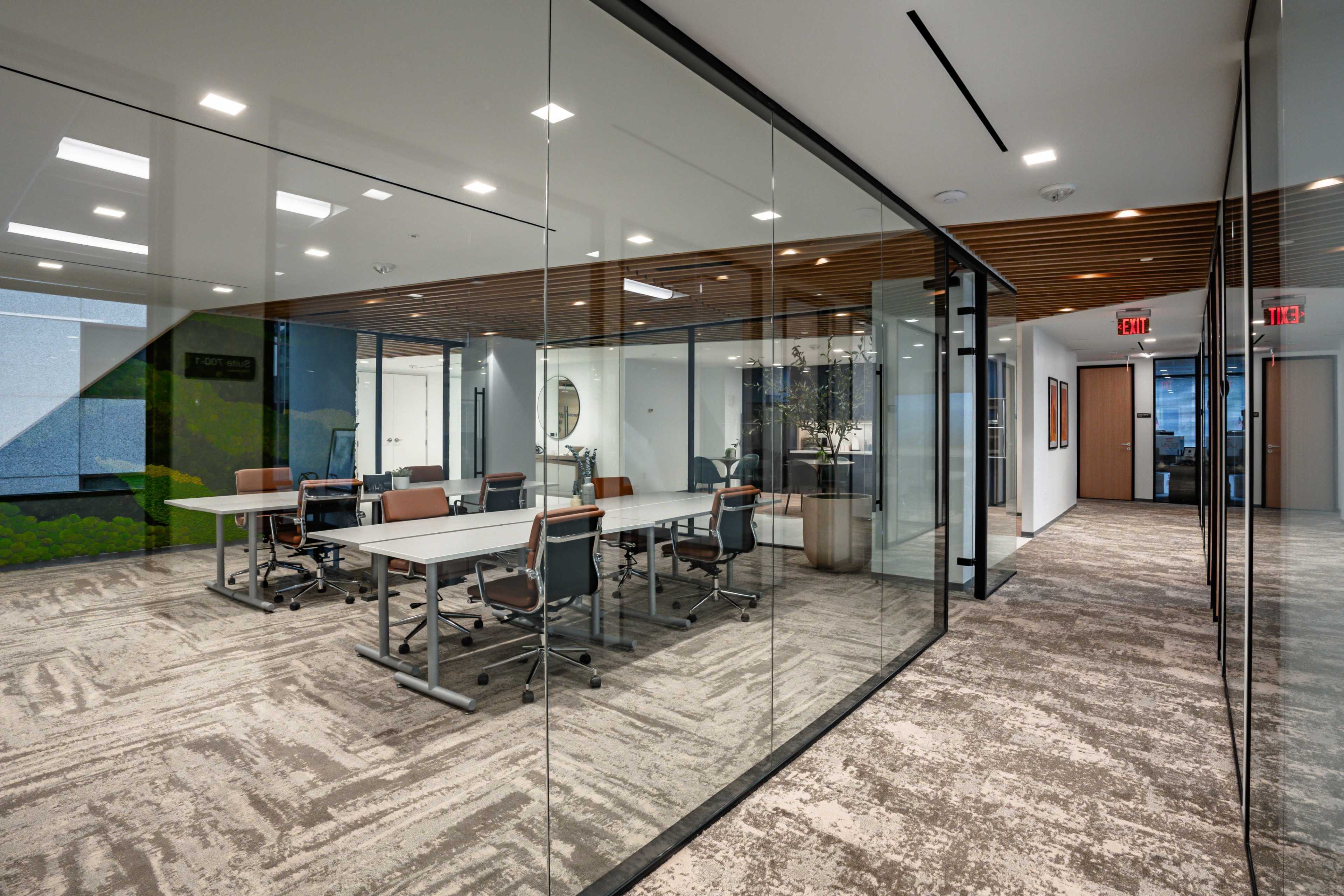 A modern office interior features a glass-walled conference room with a large table and rolling chairs, adjacent to an open hallway.