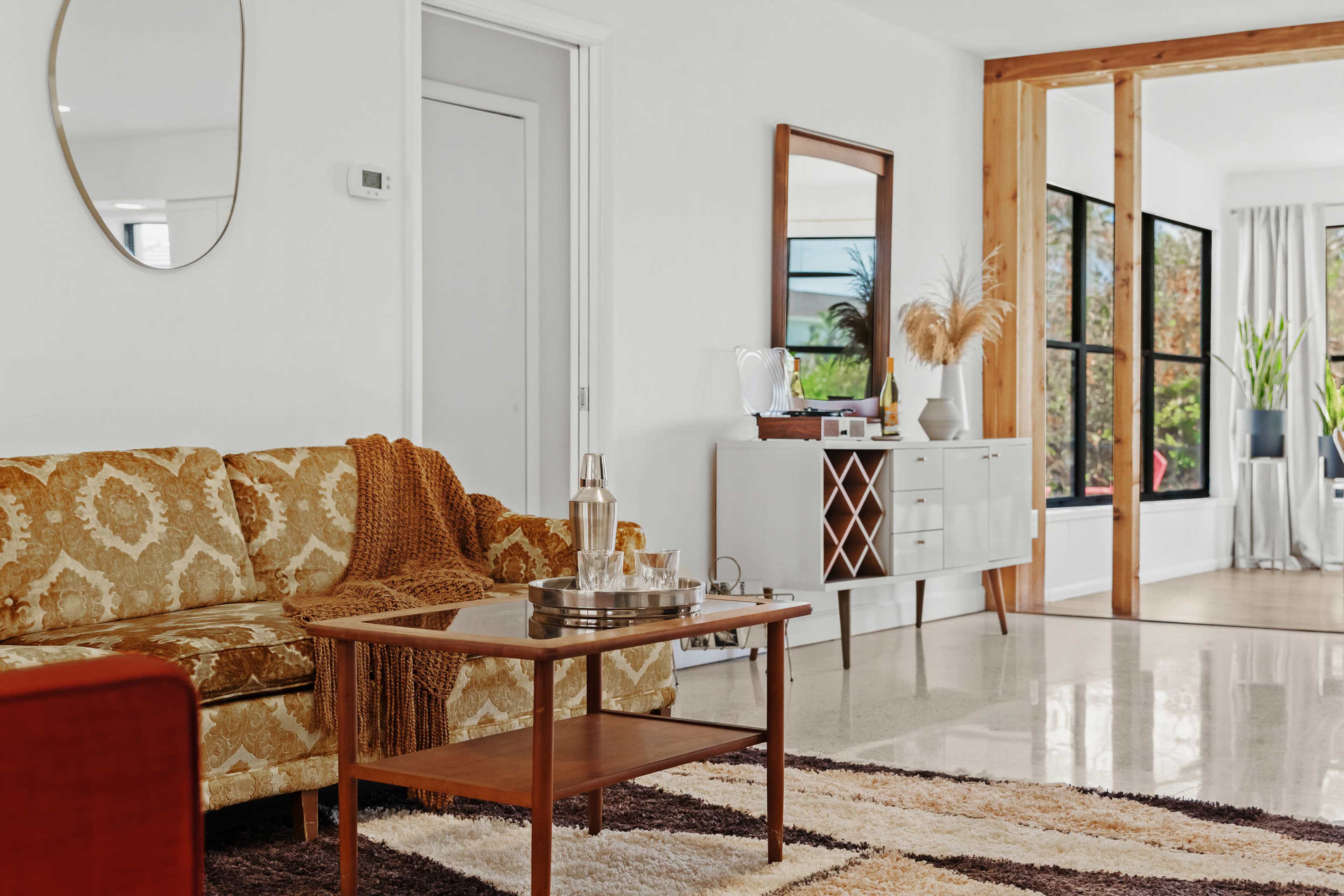 The image shows a modern living room with a patterned sofa, a coffee table set with a tray, a sideboard, and large windows allowing natural light to fill the space.