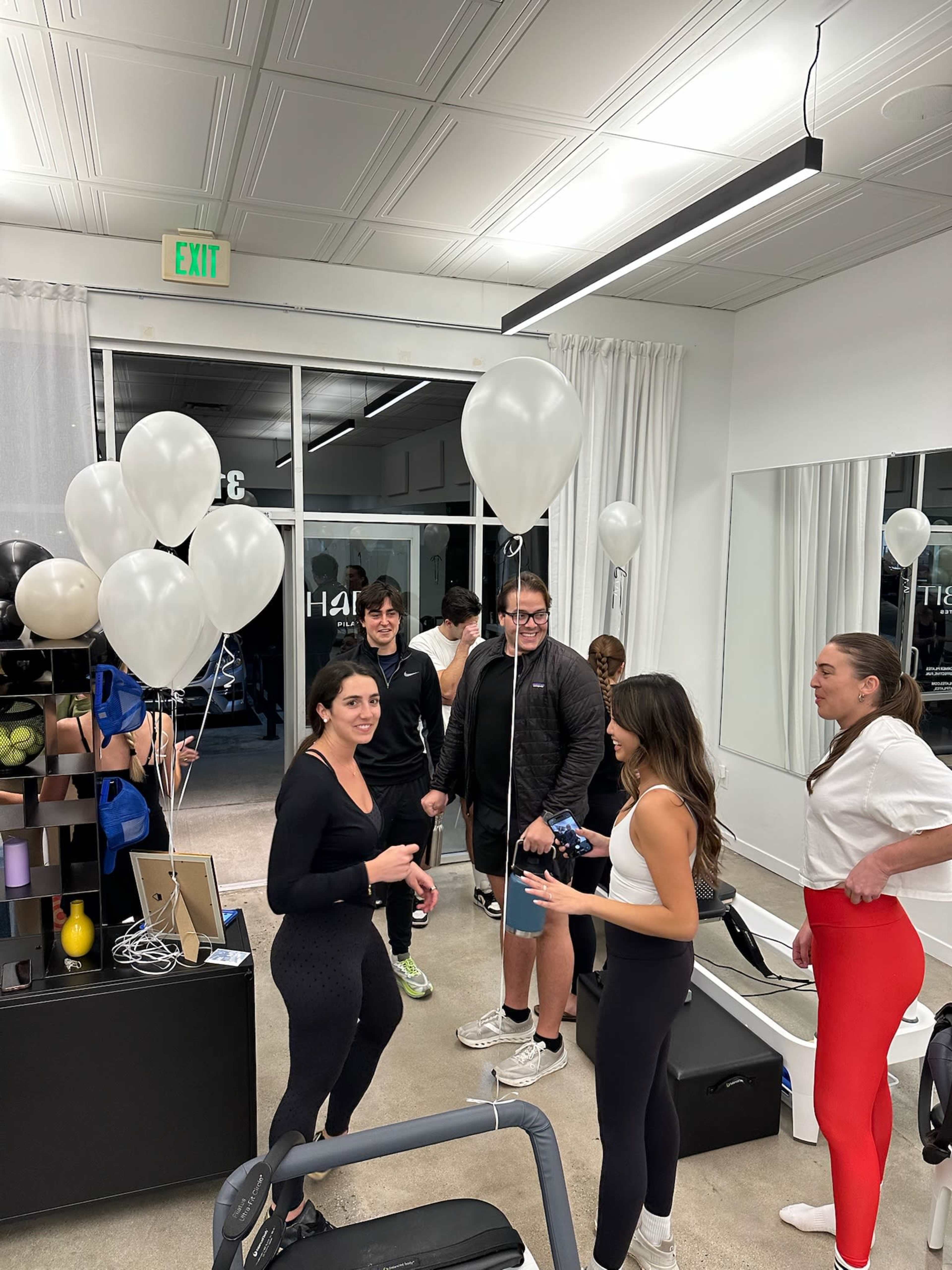 A group of five people is gathered inside a fitness studio, with balloons and exercise equipment visible in the background.