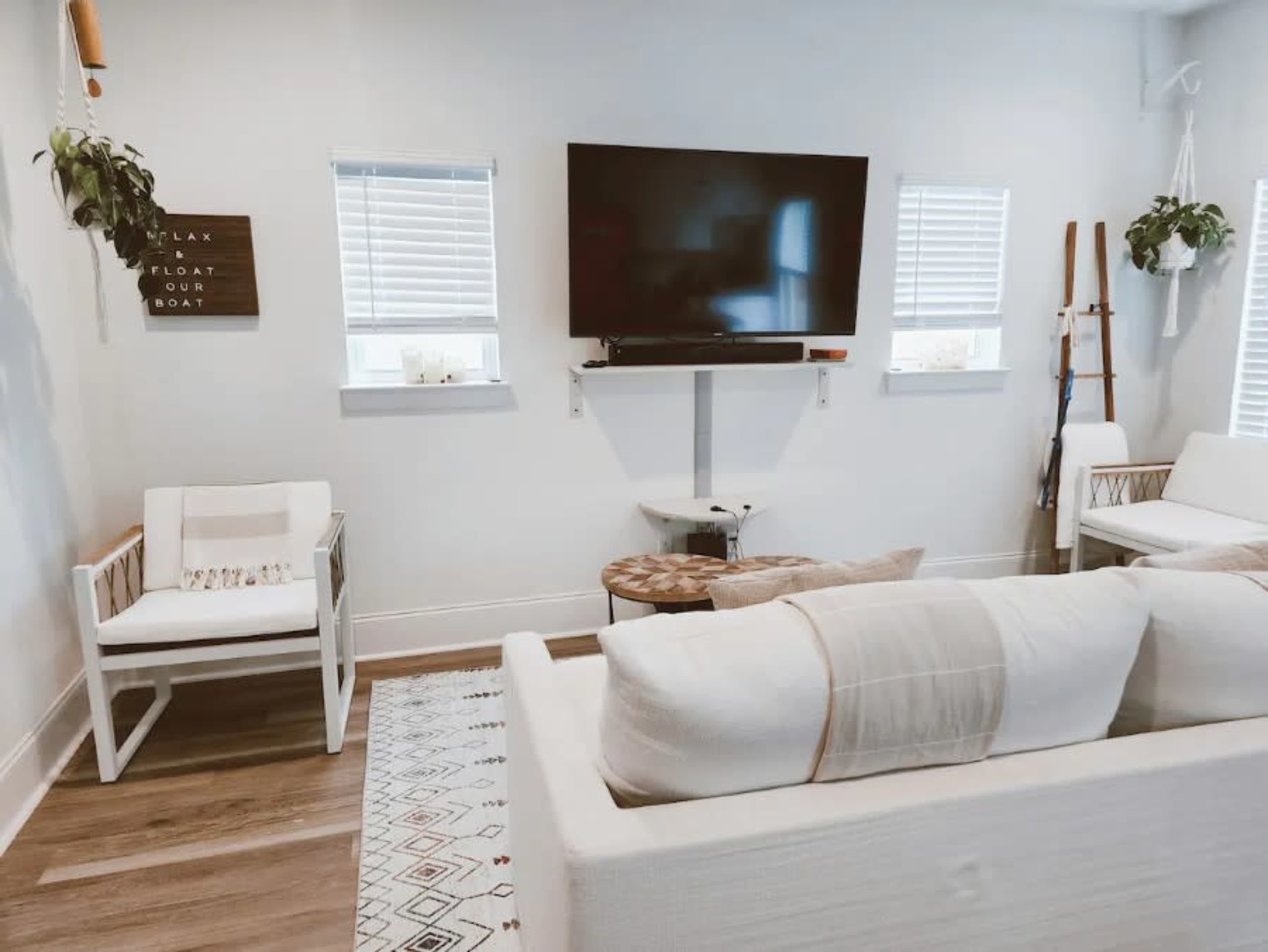 A modern living room featuring a white sofa, a television mounted on the wall, potted plants, and a patterned rug.