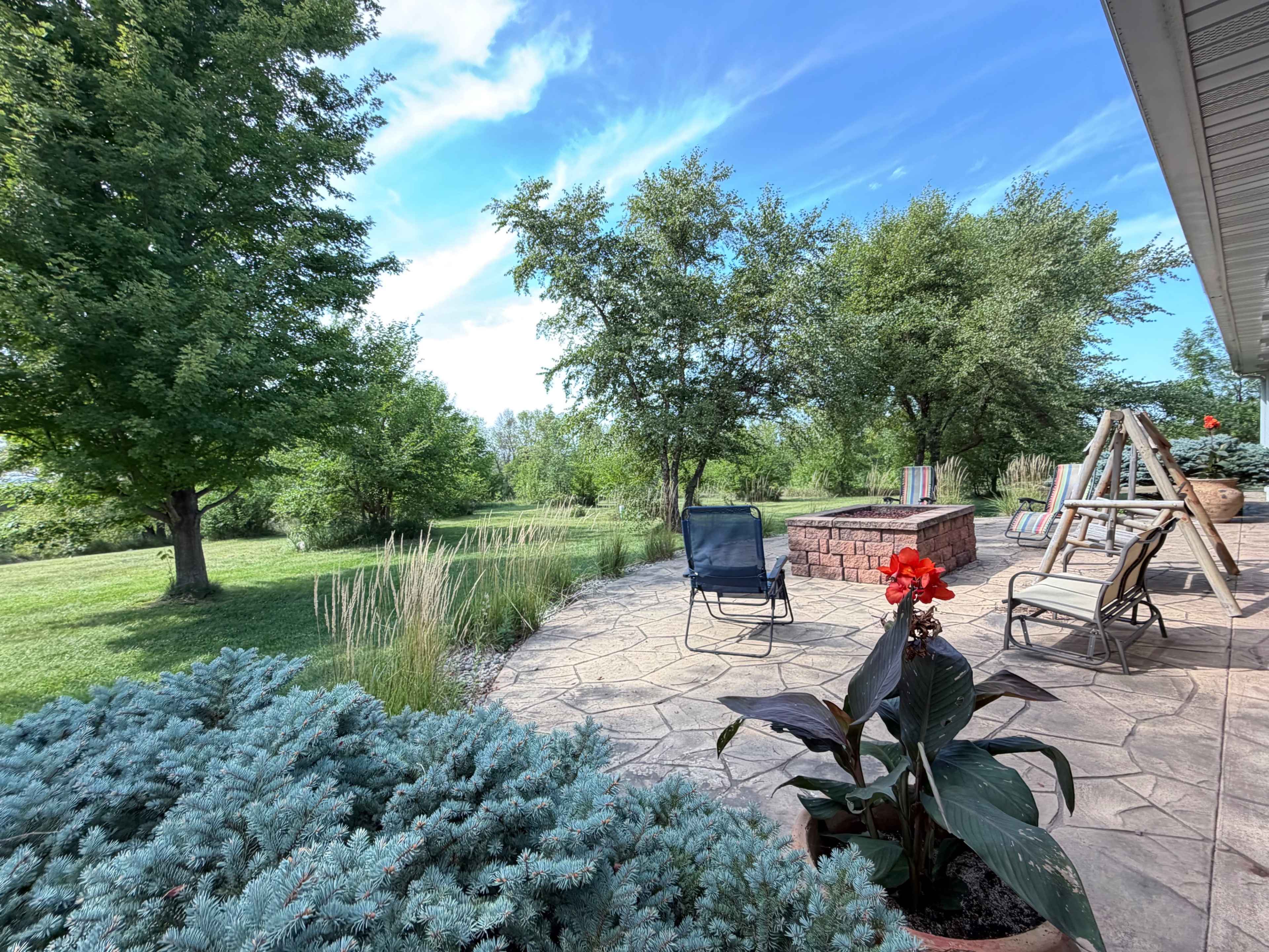 The image shows a landscaped backyard with a patio, seating area, and a fire pit surrounded by greenery and trees.