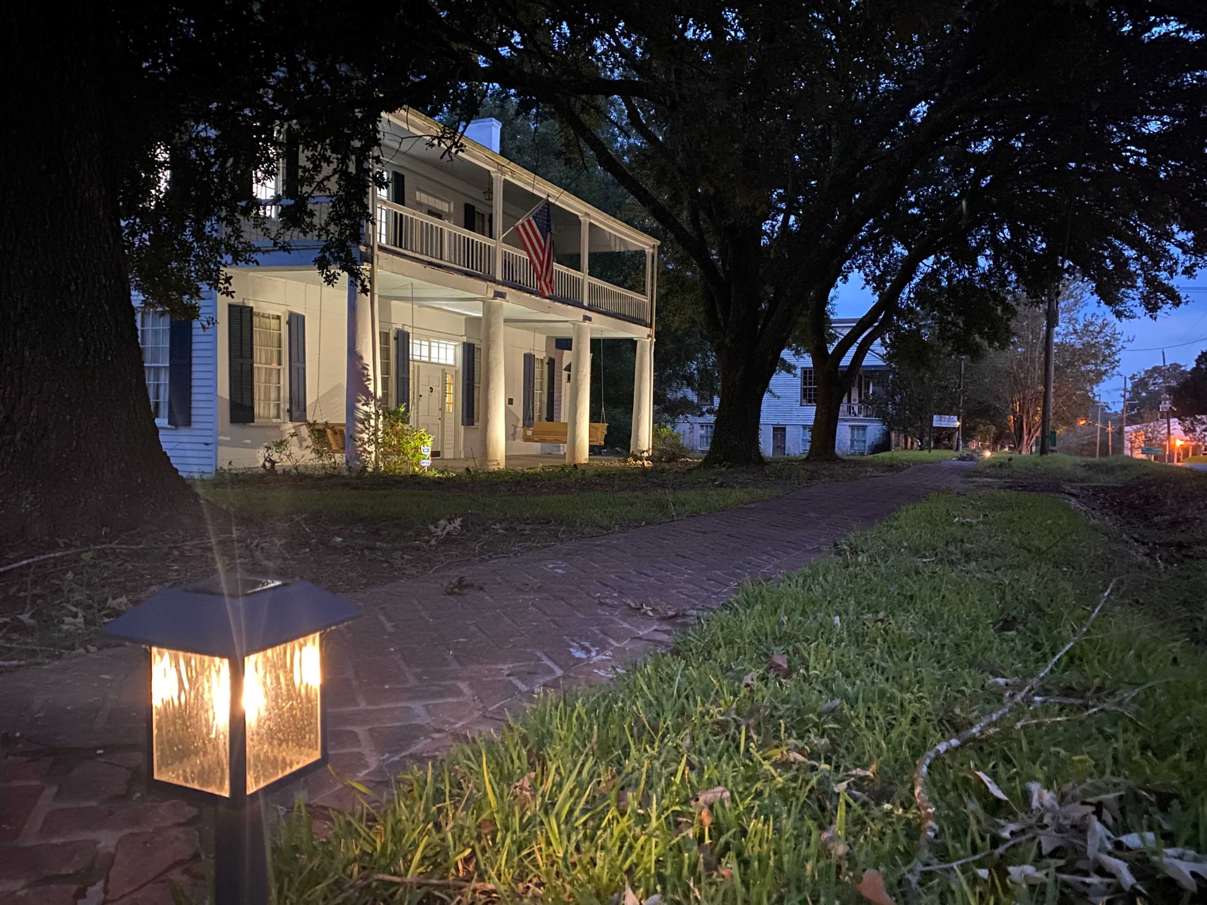 A well-lit path leads to a two-story white house with a porch and an American flag, surrounded by trees during twilight.