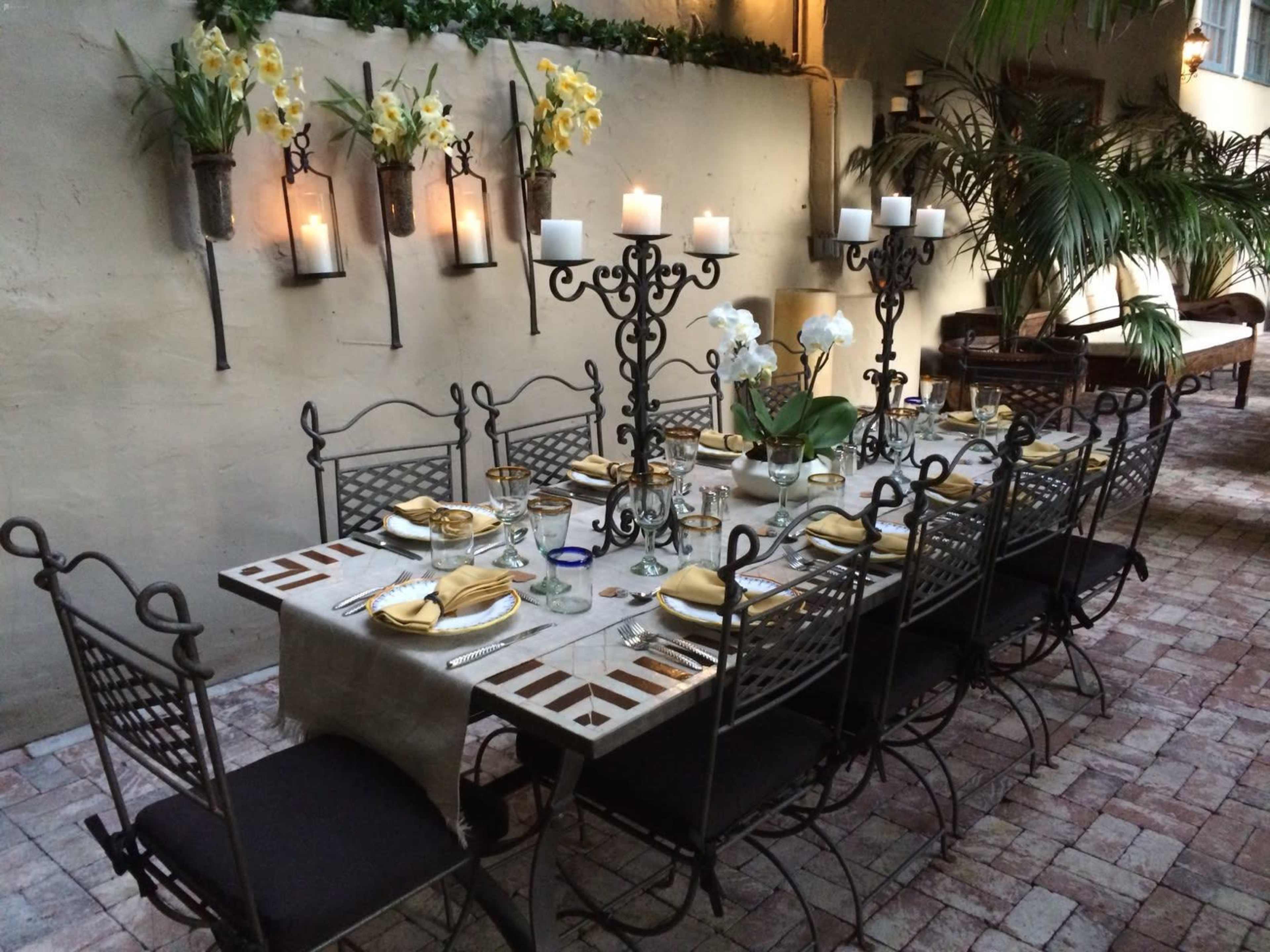 A long, decorated dining table with elegant place settings and tall candle holders is set in a courtyard surrounded by plants and walls adorned with flowers.