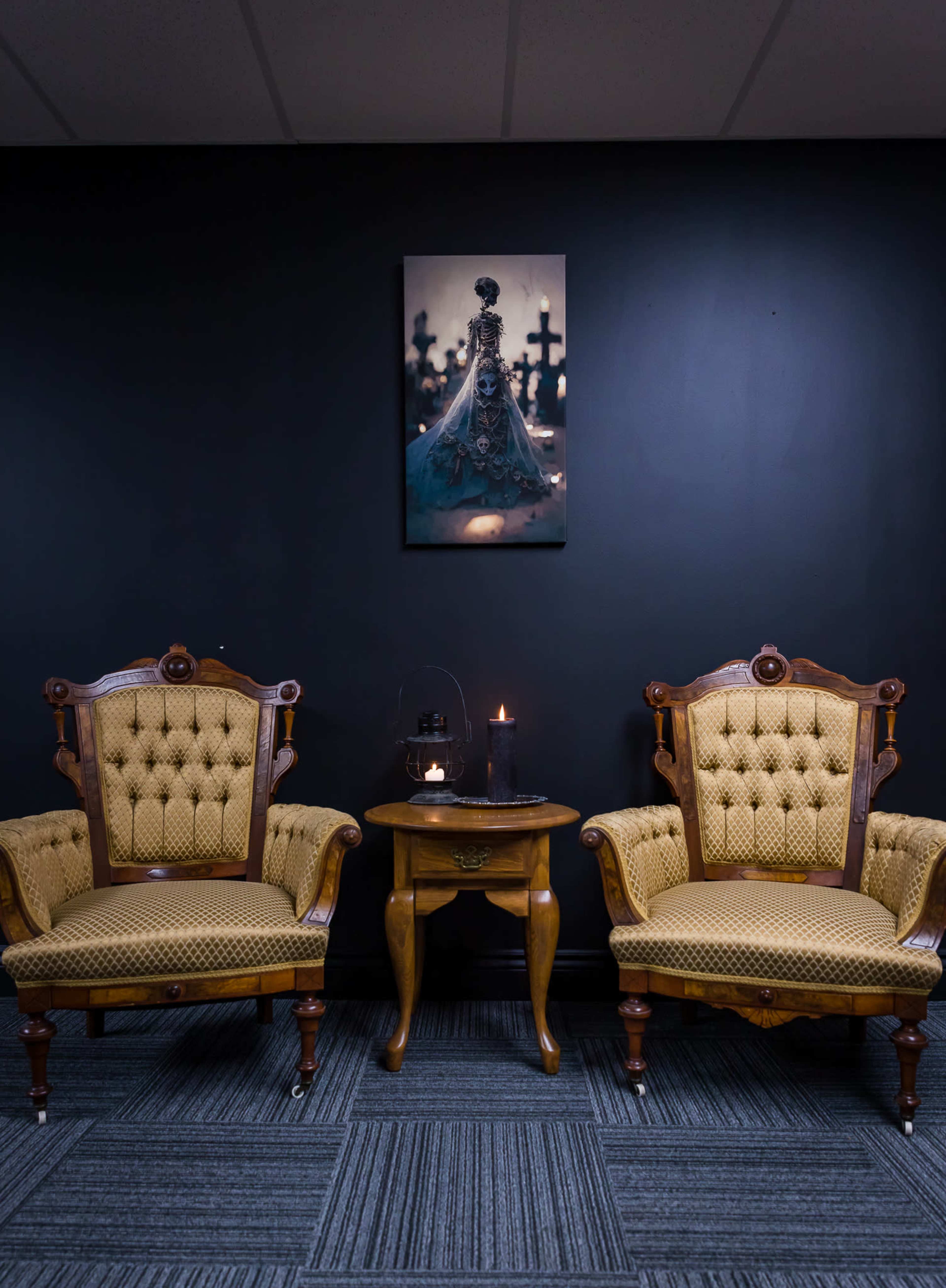 Two vintage tufted chairs flank a small wooden table against a dark wall, with a framed artwork hanging above and candles lit on the table.