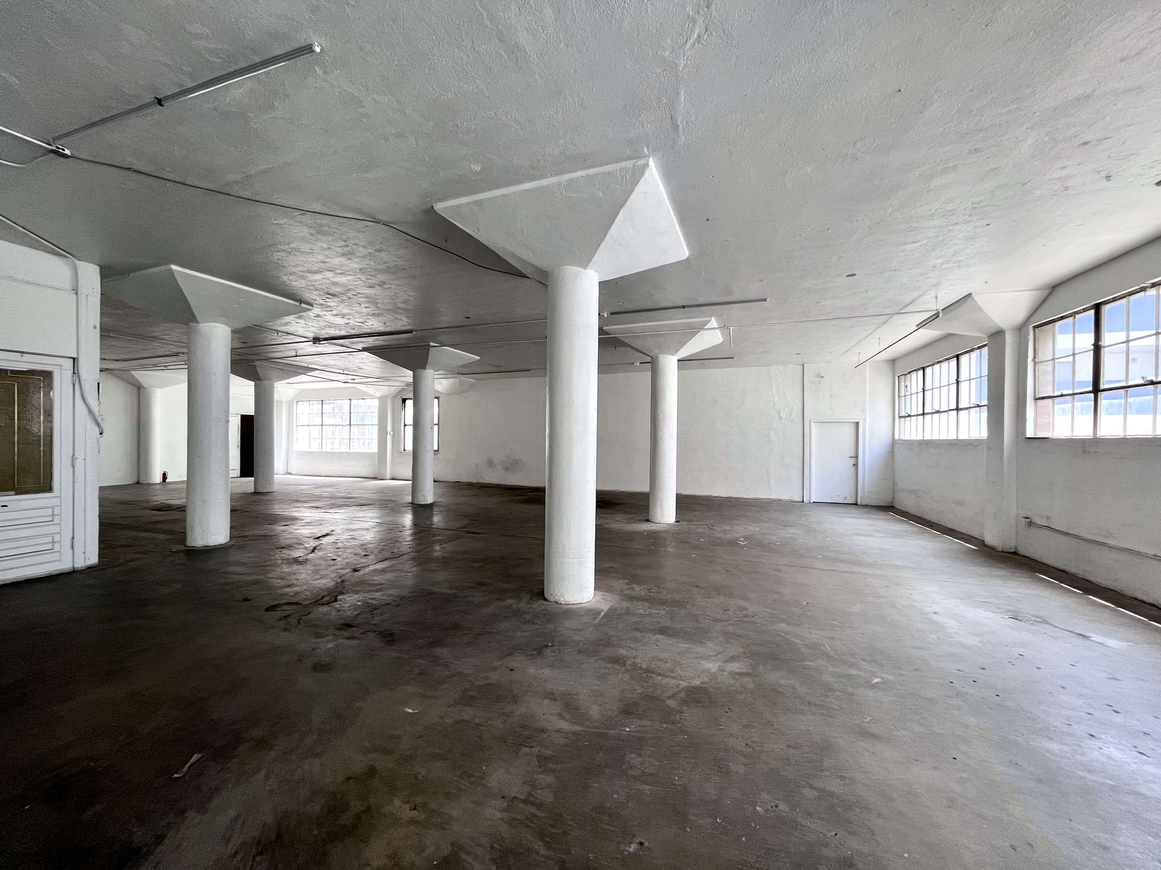A spacious, empty industrial room with white walls, large windows, and several concrete pillars.