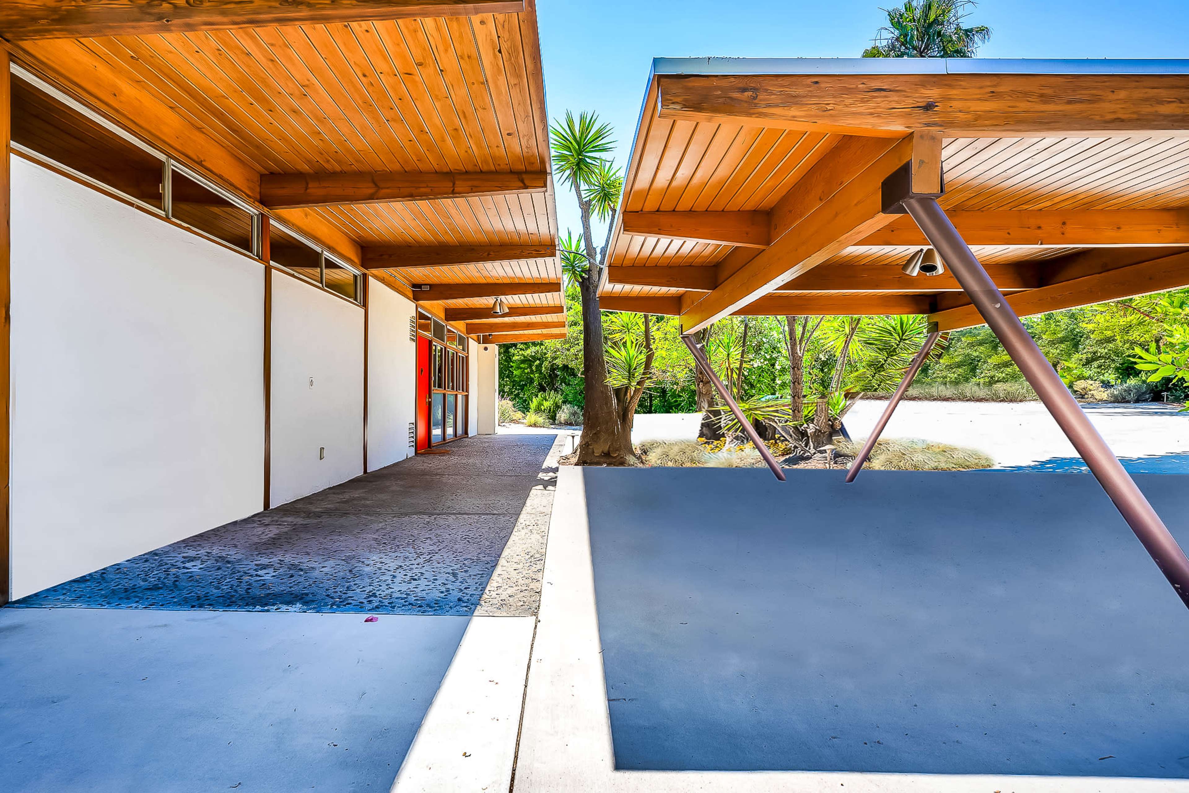 The image shows a modern architectural structure with wooden overhangs and concrete pathways, surrounded by tropical vegetation.