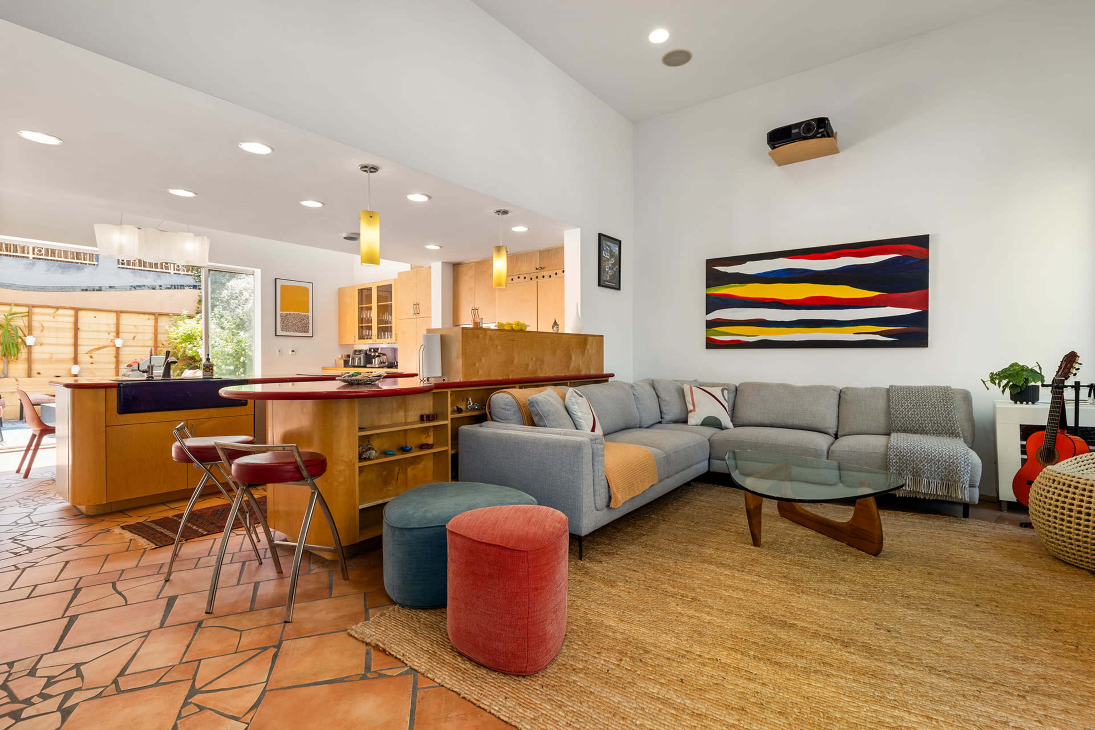 The image shows a modern living room with a sectional sofa, a glass coffee table, and an open kitchen area featuring wooden cabinetry and a colorful abstract painting on the wall.