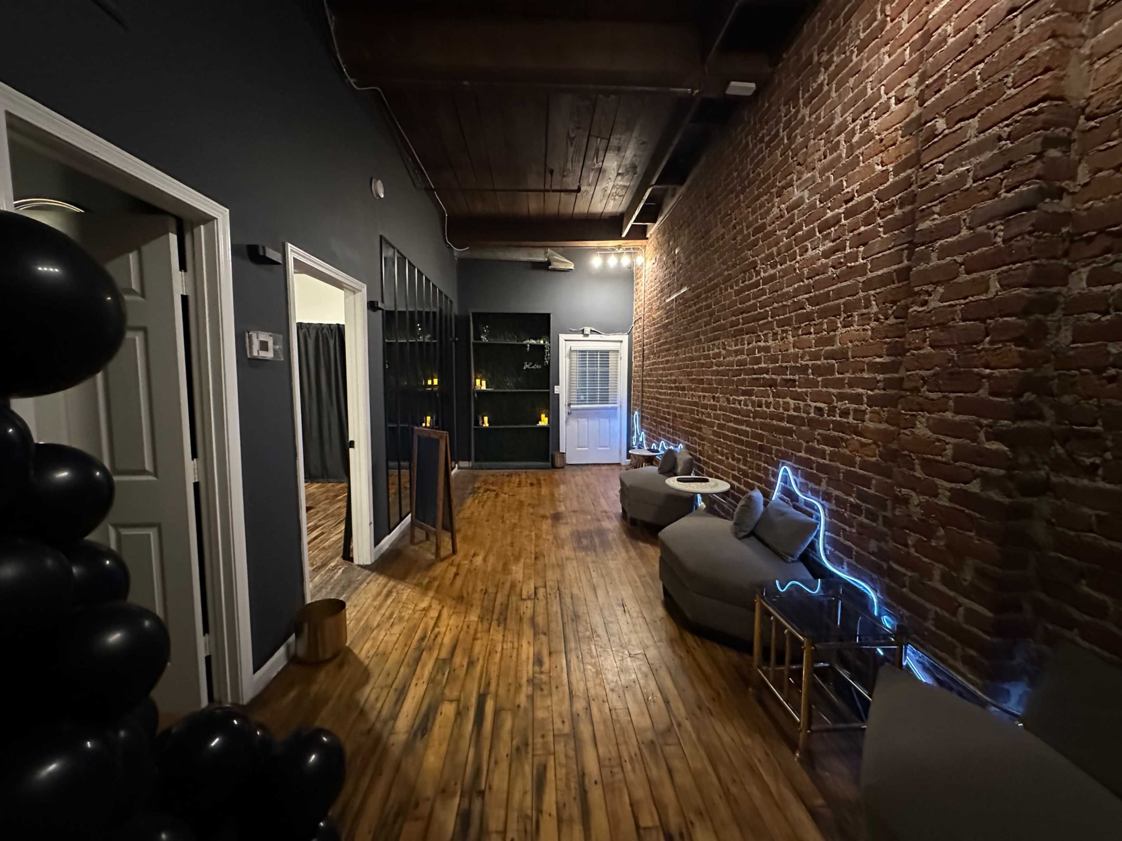 The image shows a dimly lit corridor with exposed brick walls, wooden flooring, and seating areas featuring gray cushions along one side.