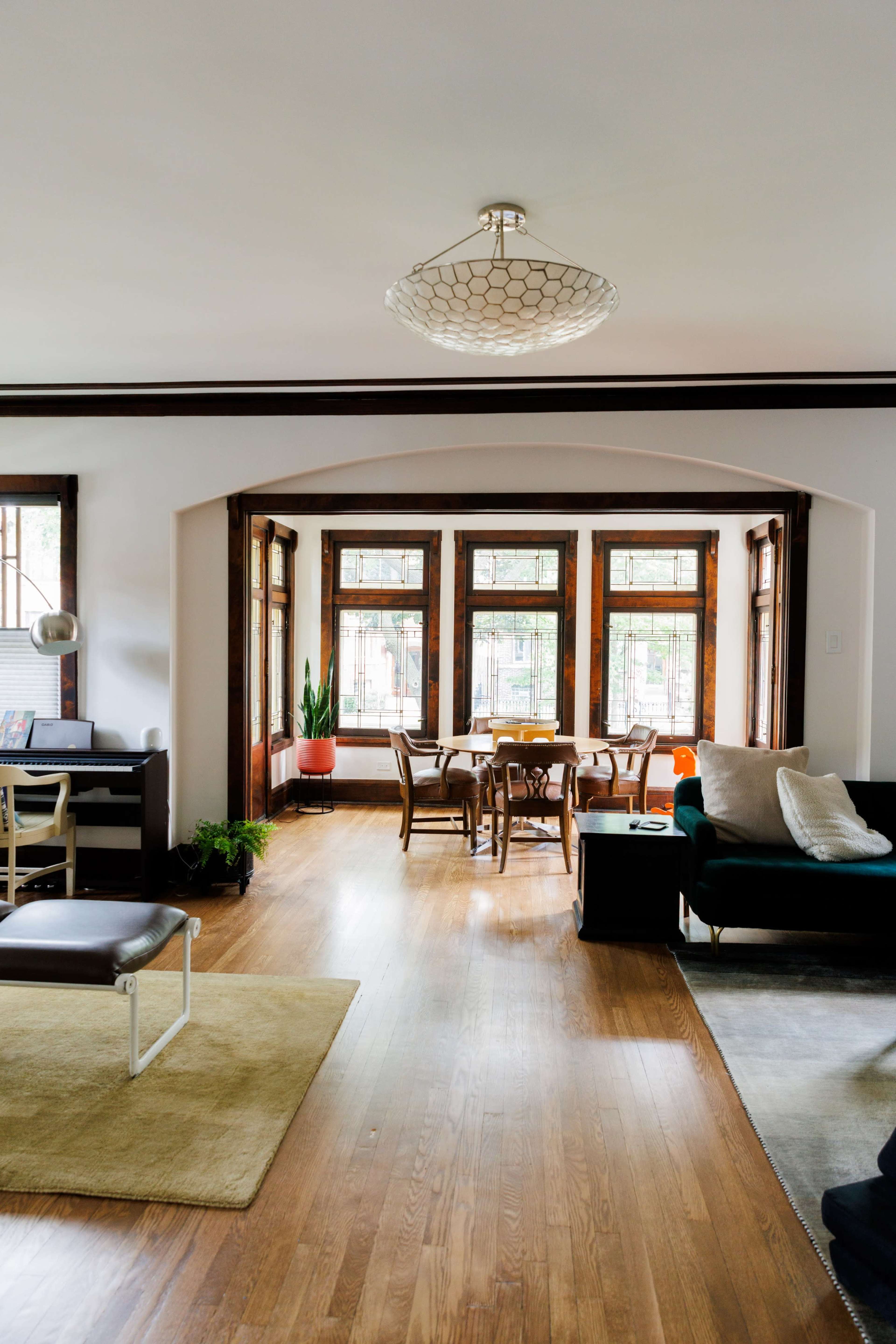 A spacious living area features a modern chandelier, wooden floors, and large windows with a dining table in view.