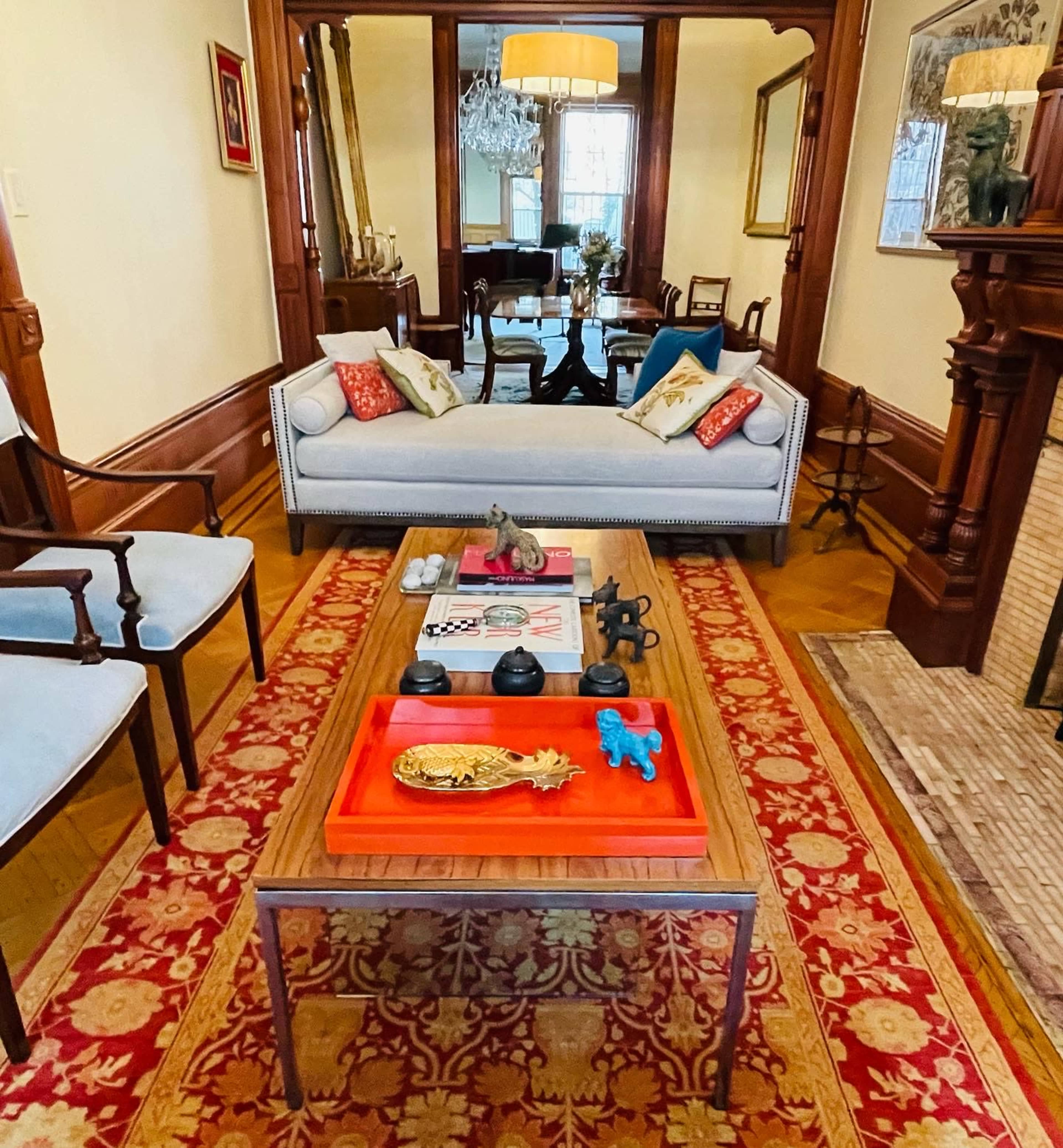 A living room with a decorative coffee table center, flanked by two chairs, and featuring a traditional rug, wood paneling, and a chandelier in the background.