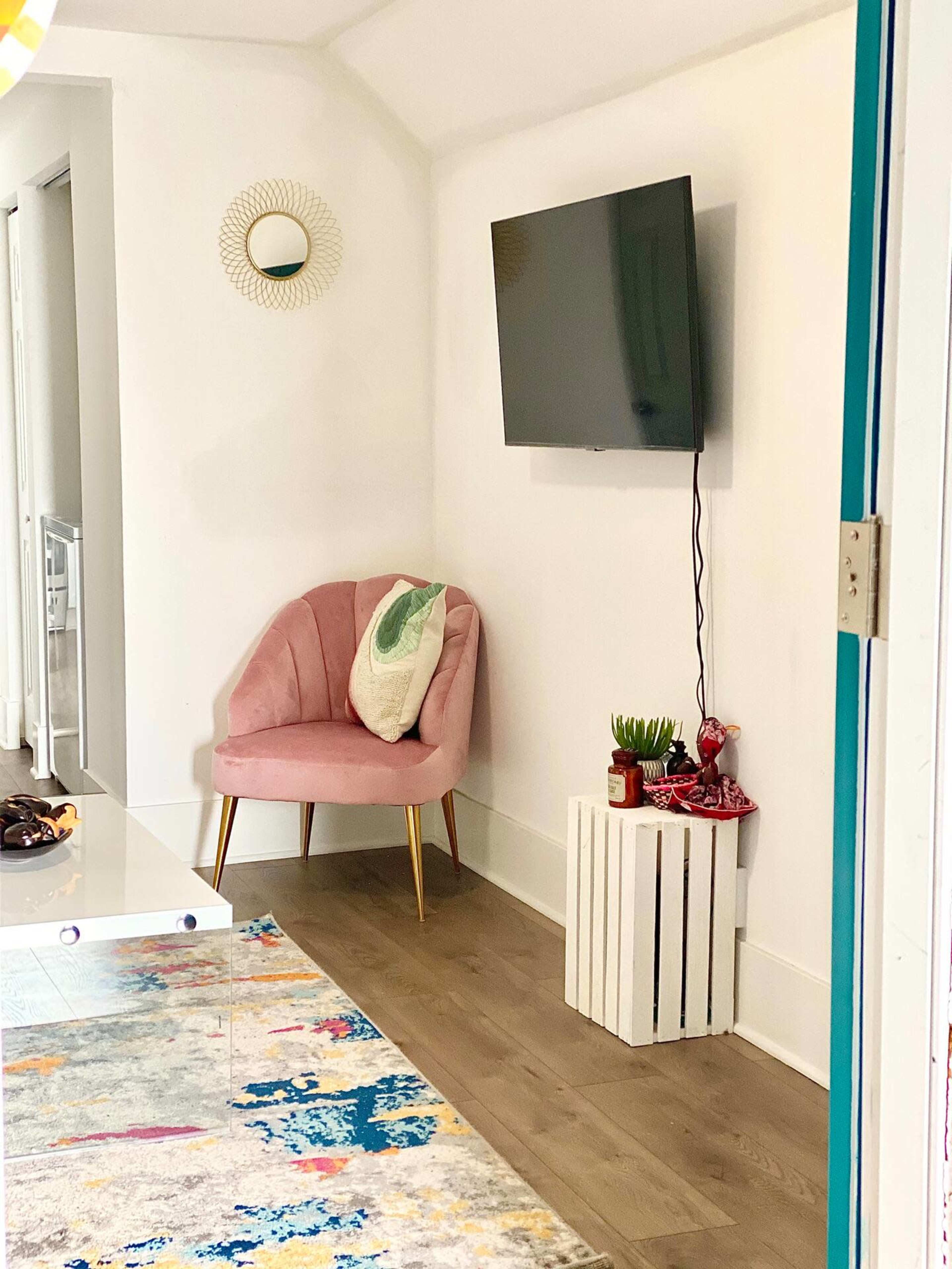 The image shows a small corner of a room featuring a pink chair, a wall-mounted television, a white wooden crate with plants, and a colorful rug on the floor.