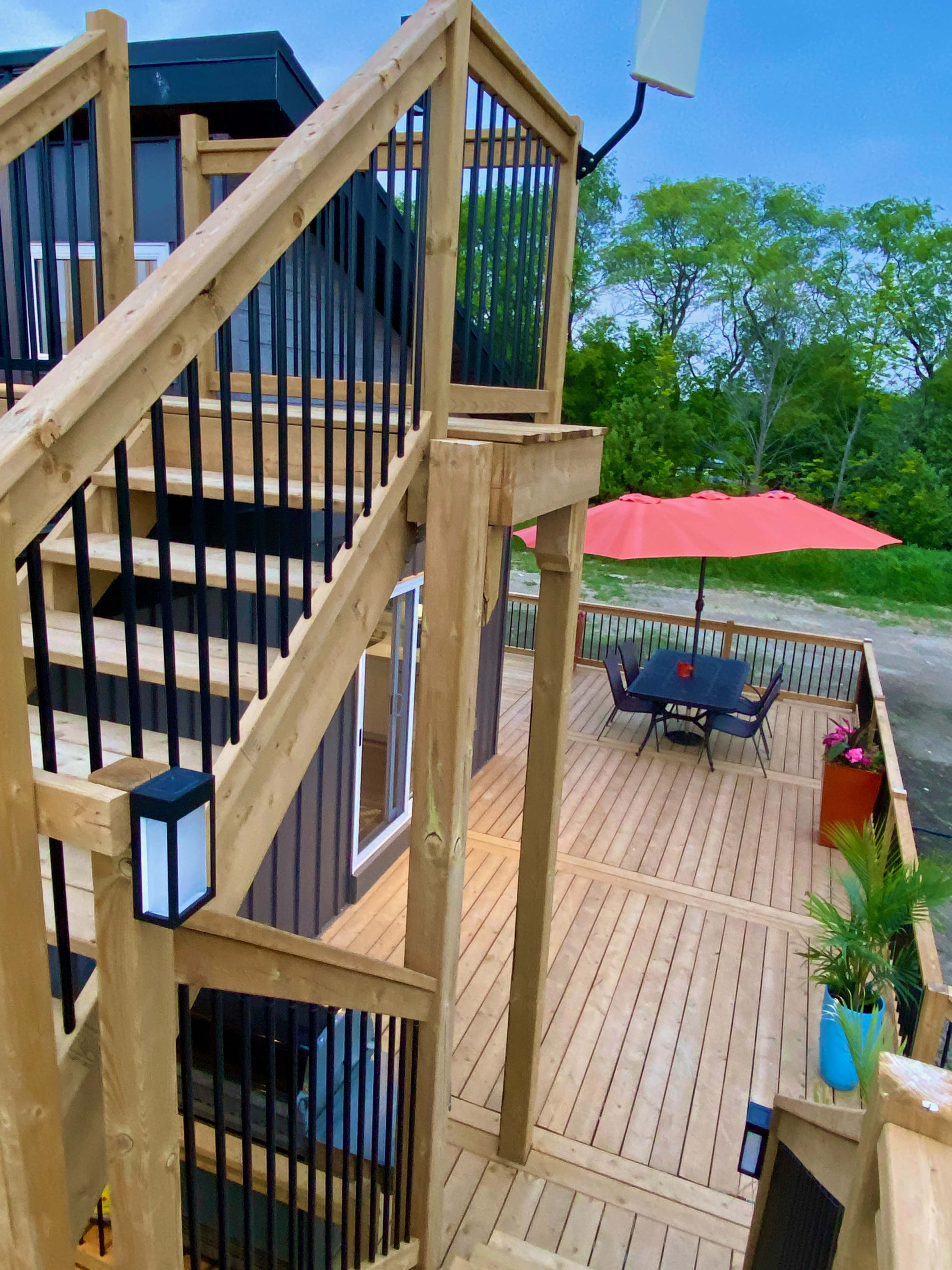 A wooden deck with a staircase, an outdoor dining area under an umbrella, and a landscaped backdrop of trees.