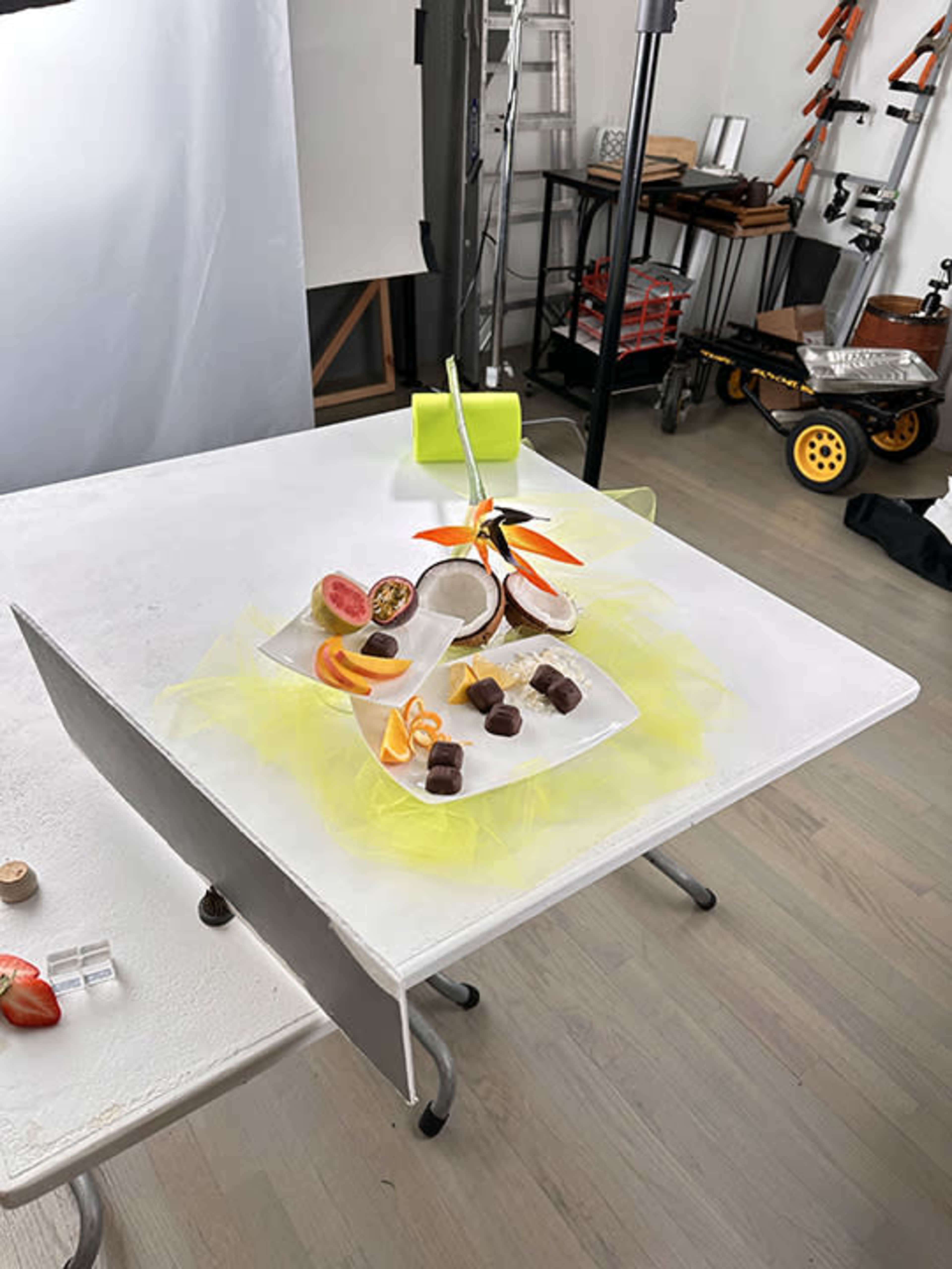 A table is set up in a photography studio with a spread of assorted fruits and chocolates, surrounded by colorful props and equipment.