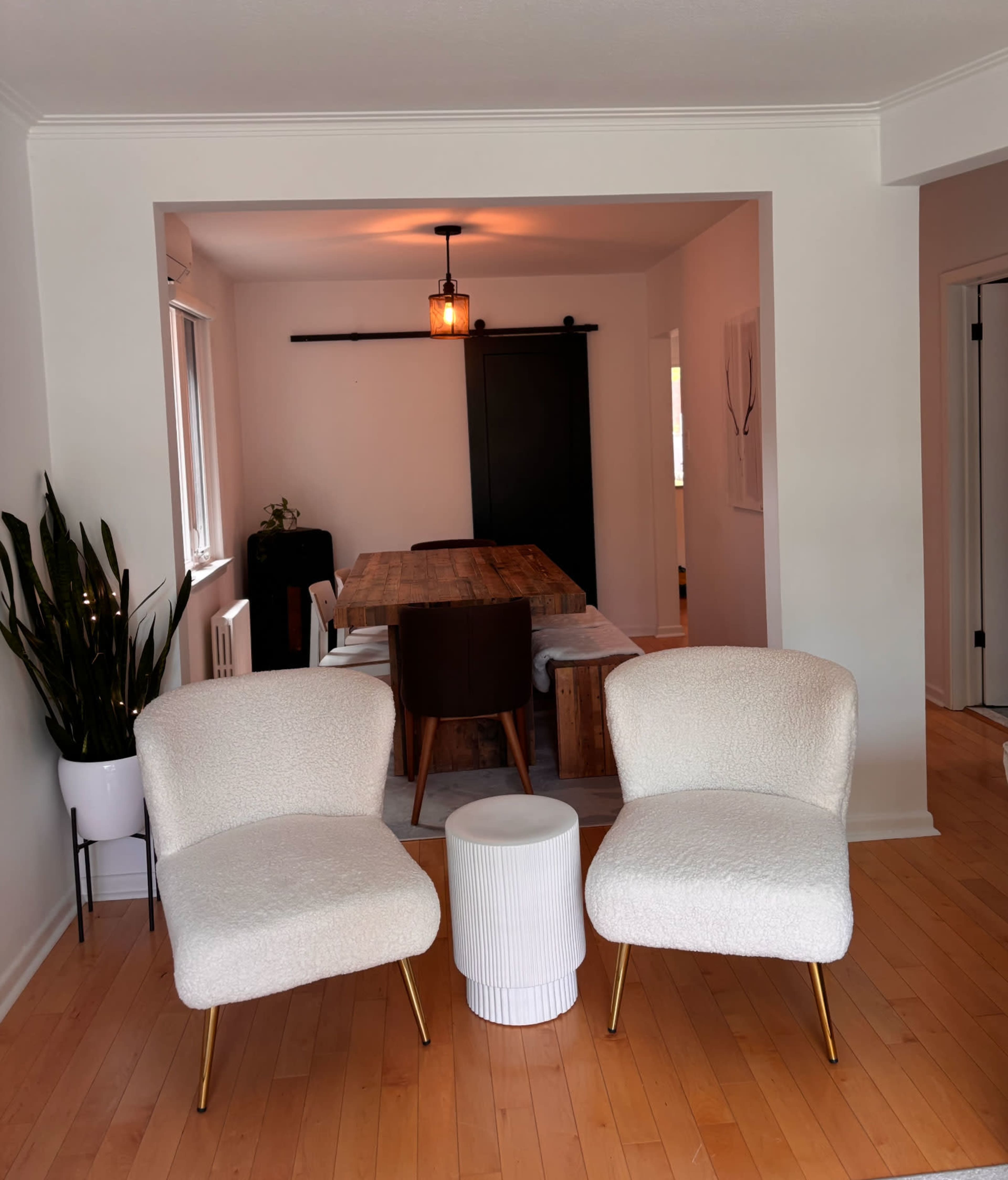 Two white, textured chairs face a small round table in a bright, open living space that leads to a dining area with a wooden table.