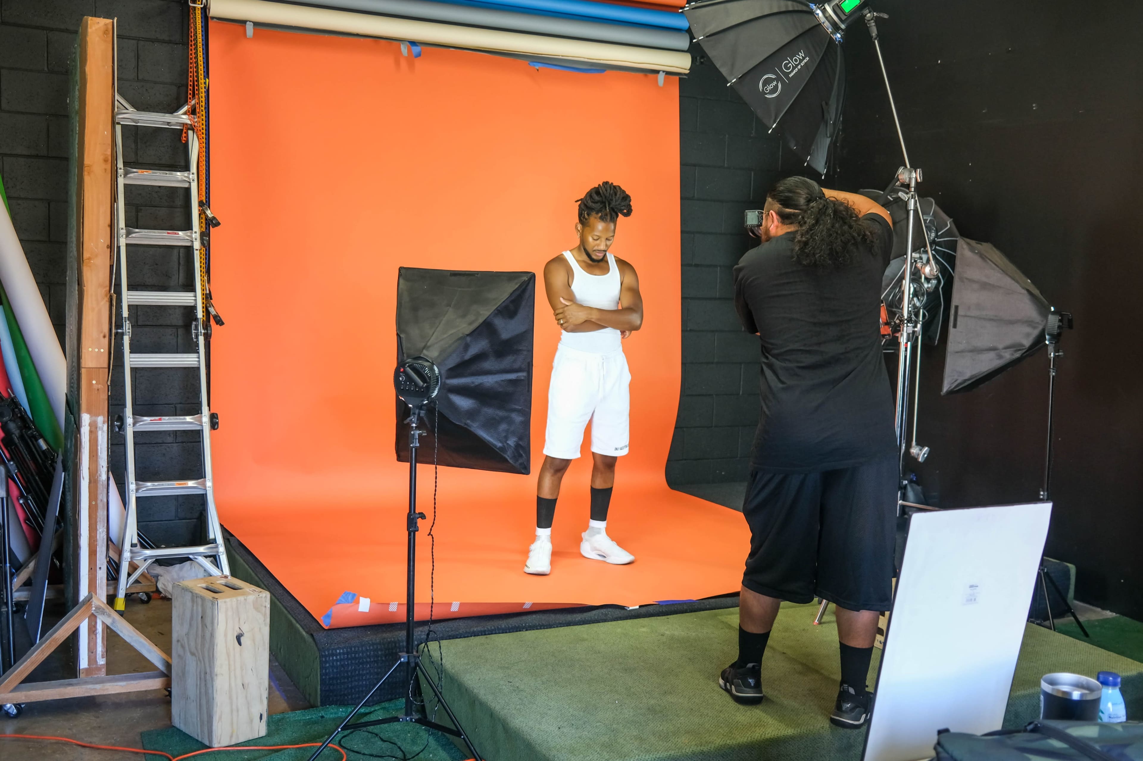 A person poses in a studio setting with an orange backdrop while a photographer adjusts the lighting and equipment.