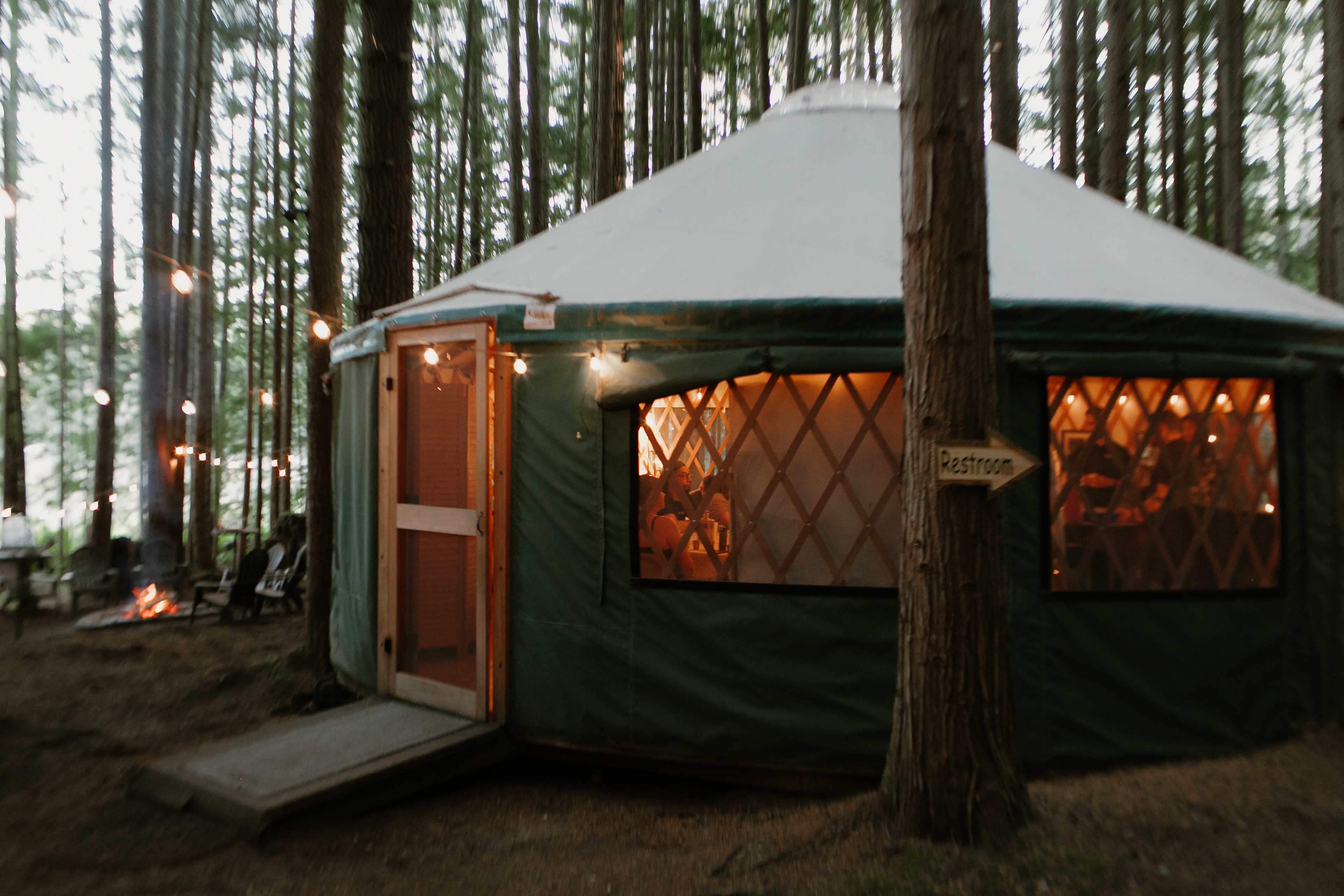 A green yurt with a wooden entrance is situated among tall trees, illuminated by soft lights, with a sign indicating the restroom nearby.
