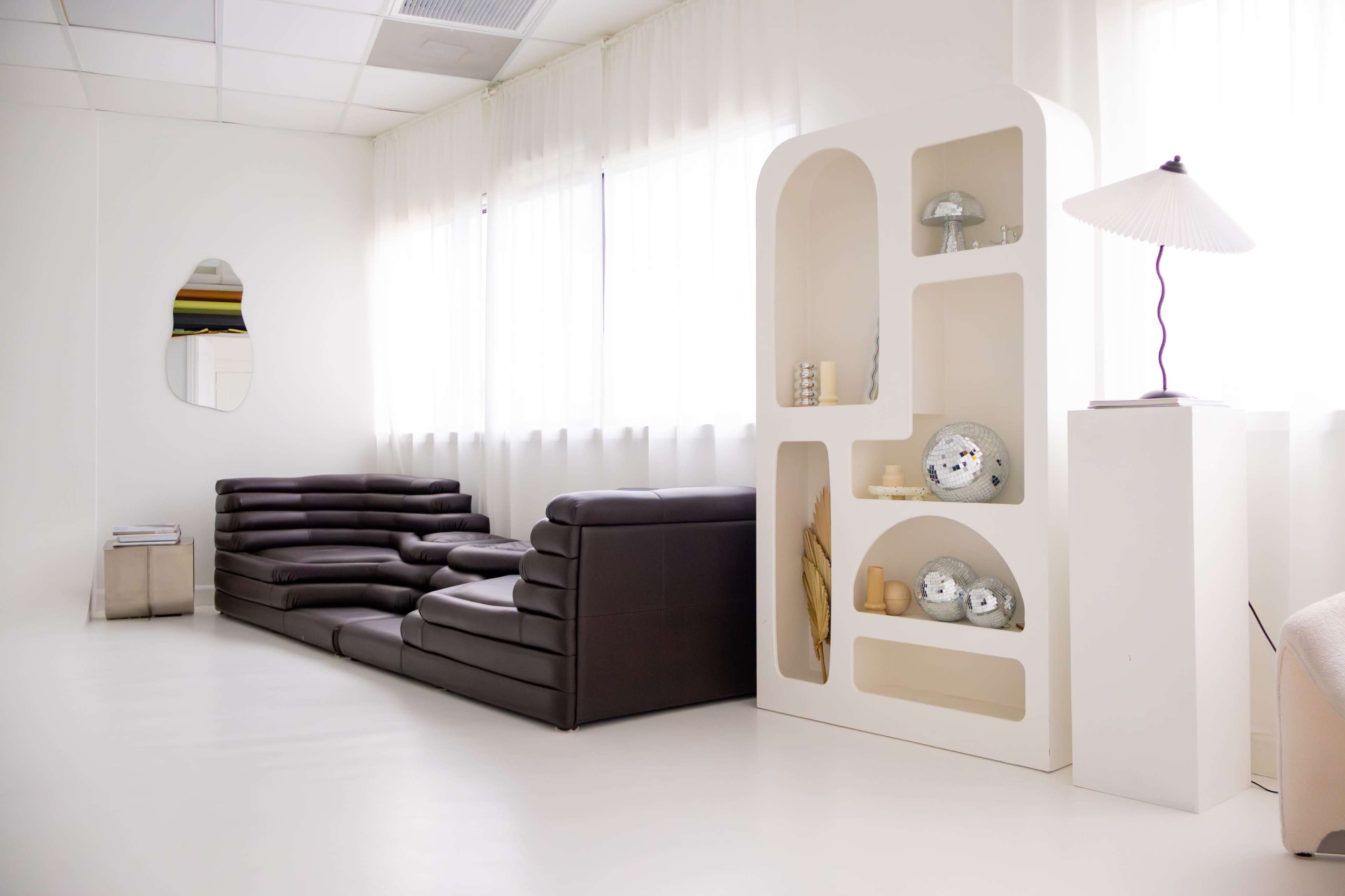 The image features a modern, minimalist interior with a dark brown sectional sofa, a white shelf displaying decorative items, and large windows allowing natural light.