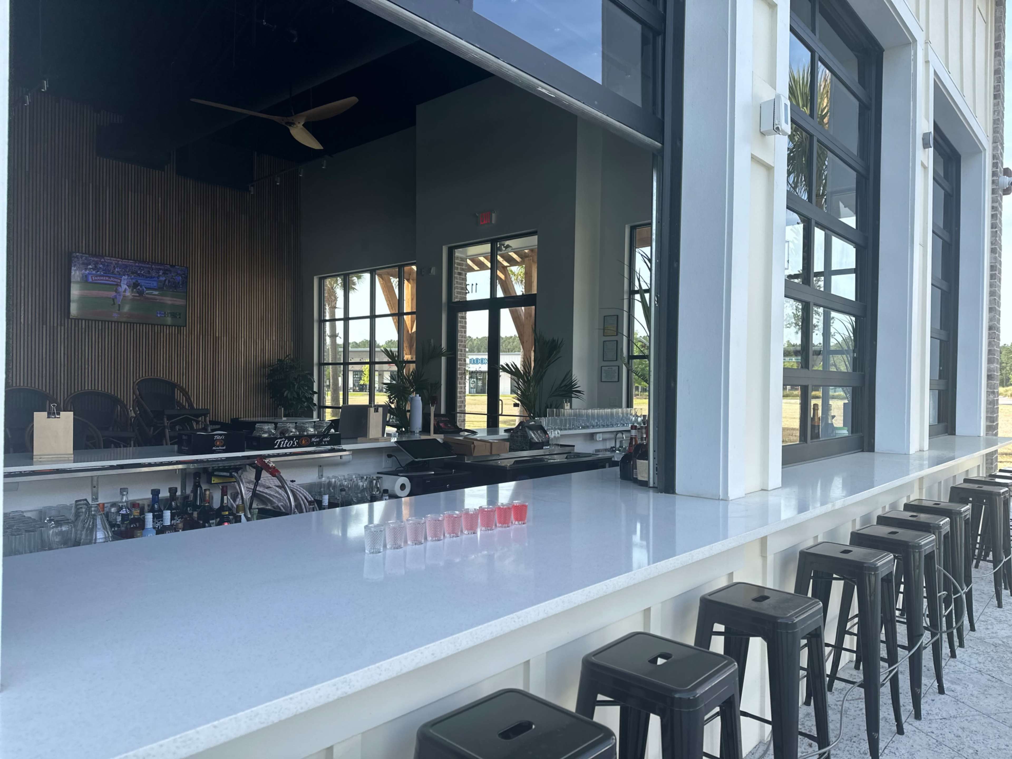The image shows a modern bar area with a white countertop, black stools, and large windows that provide a view of an outdoor space.