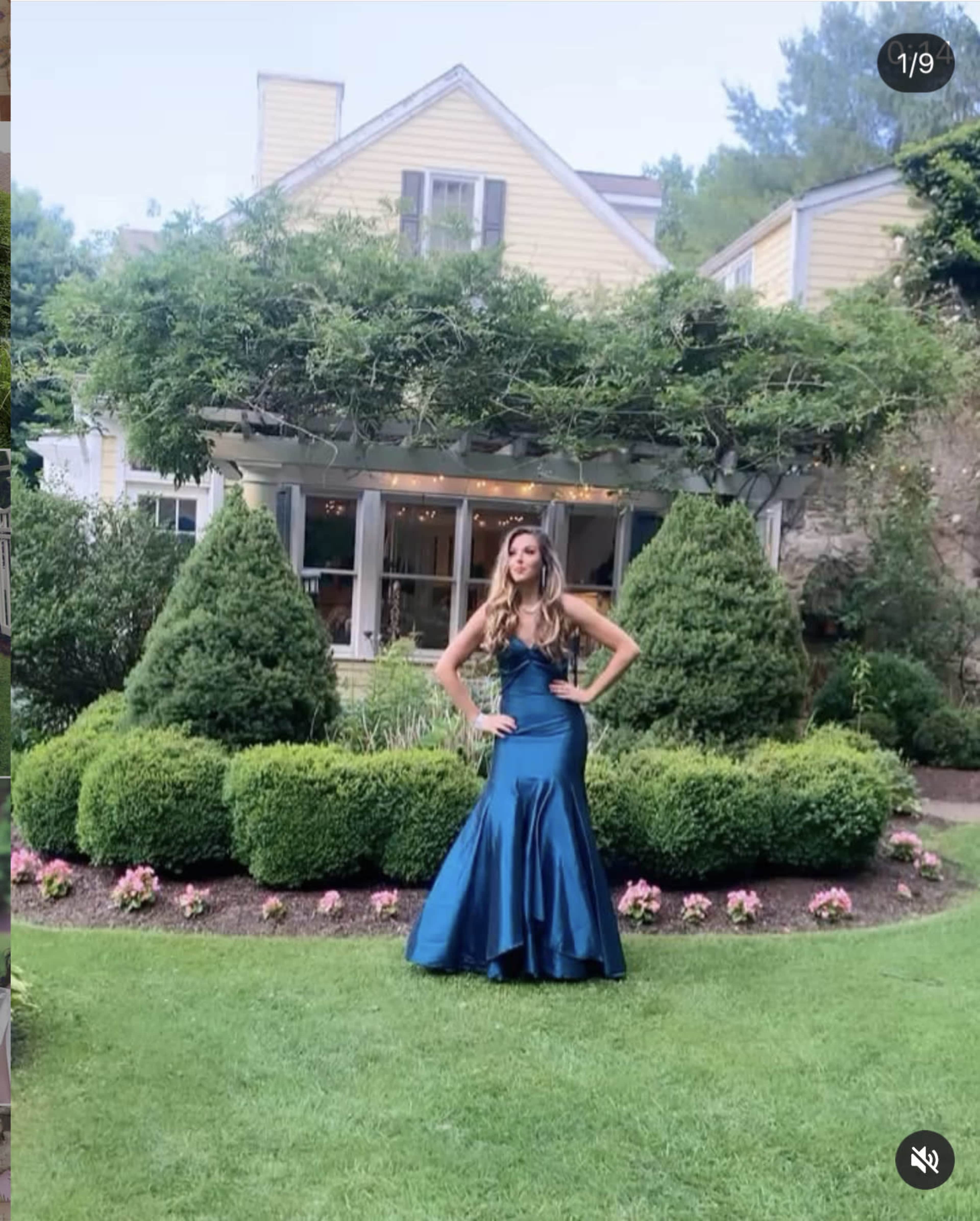 A woman in a blue gown poses in front of a house with landscaped gardens and topiary bushes.