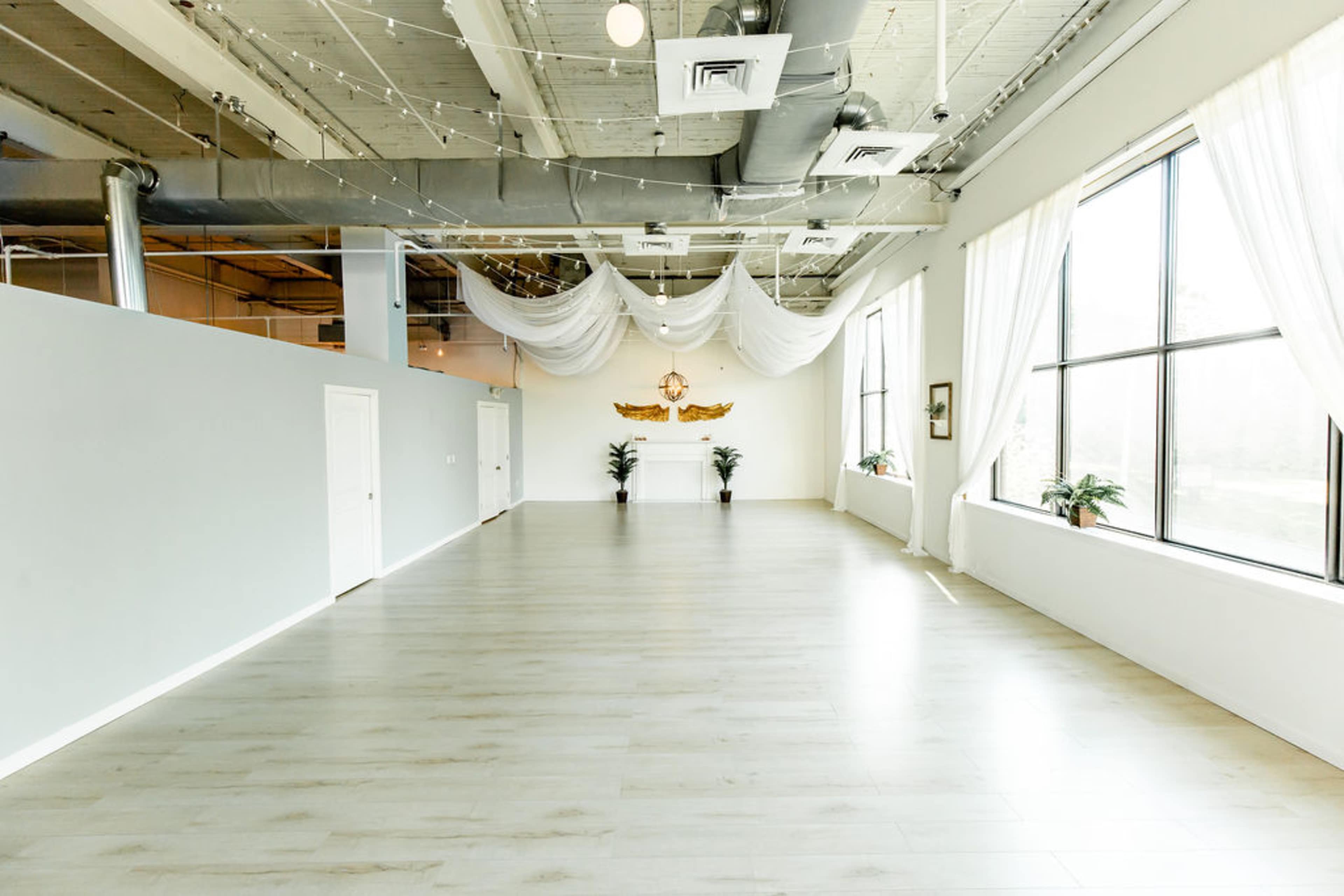 A spacious, well-lit room with large windows, light-colored wooden flooring, and white drapes hanging from the ceiling.