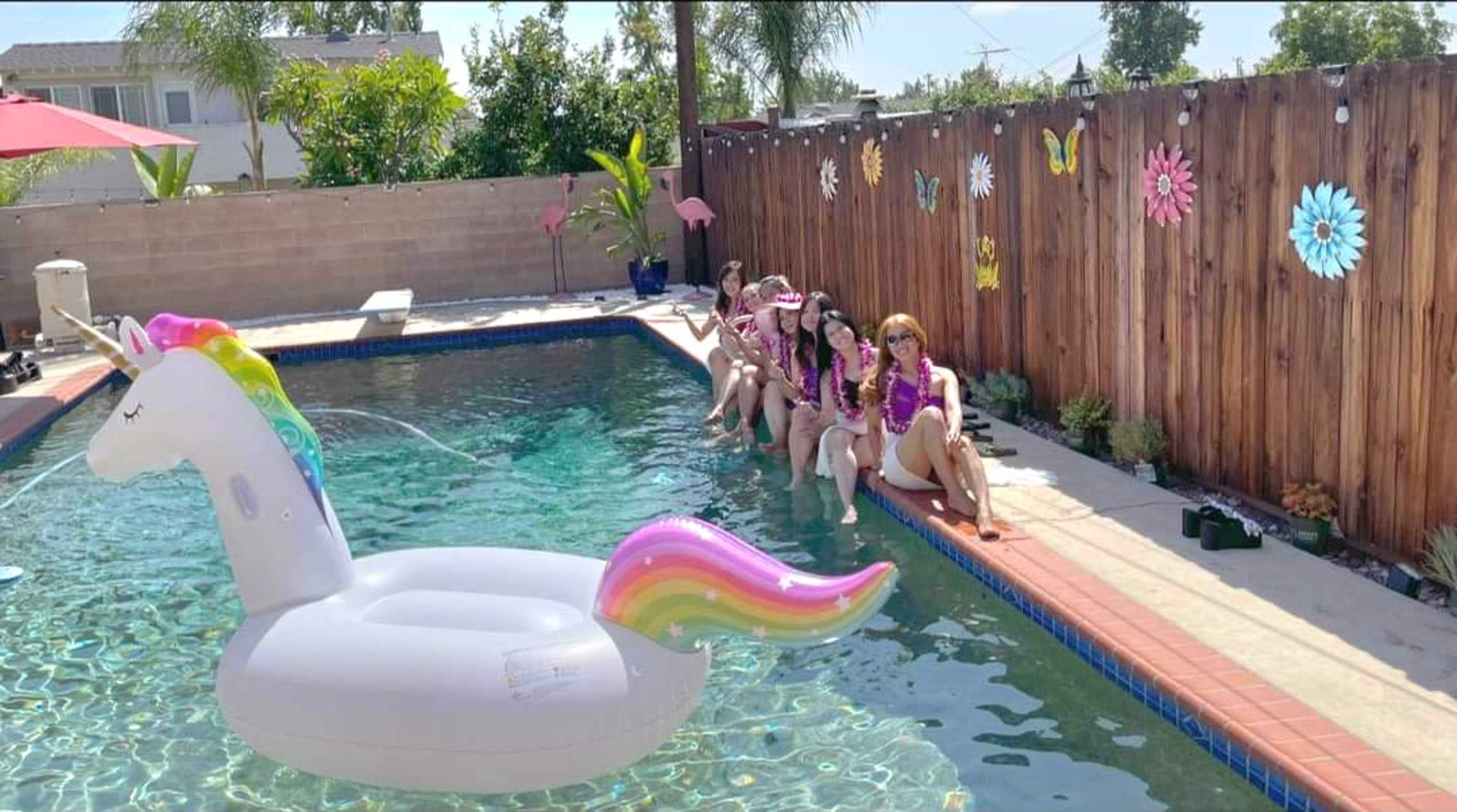A group of five people dressed in colorful attire relaxes by a swimming pool beside a large inflatable unicorn.