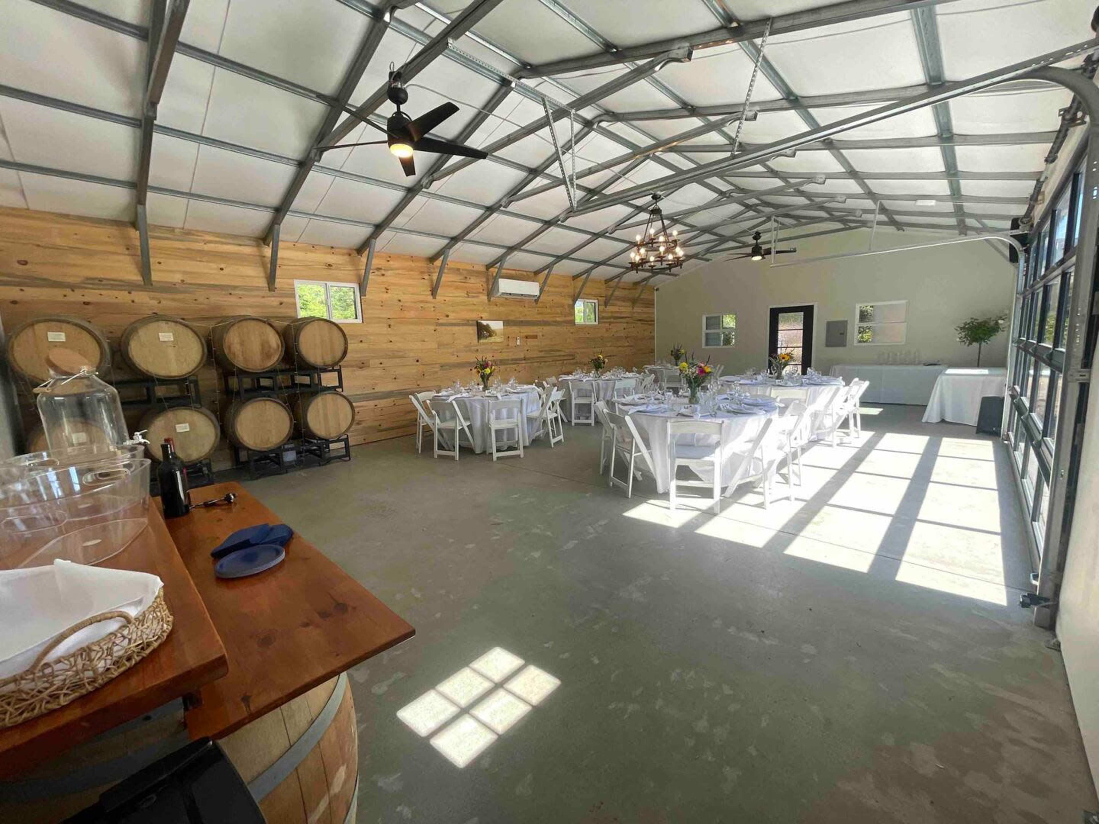 An indoor event space with white tables set for dining, wooden barrels stacked against a wall, and large windows allowing natural light to enter.