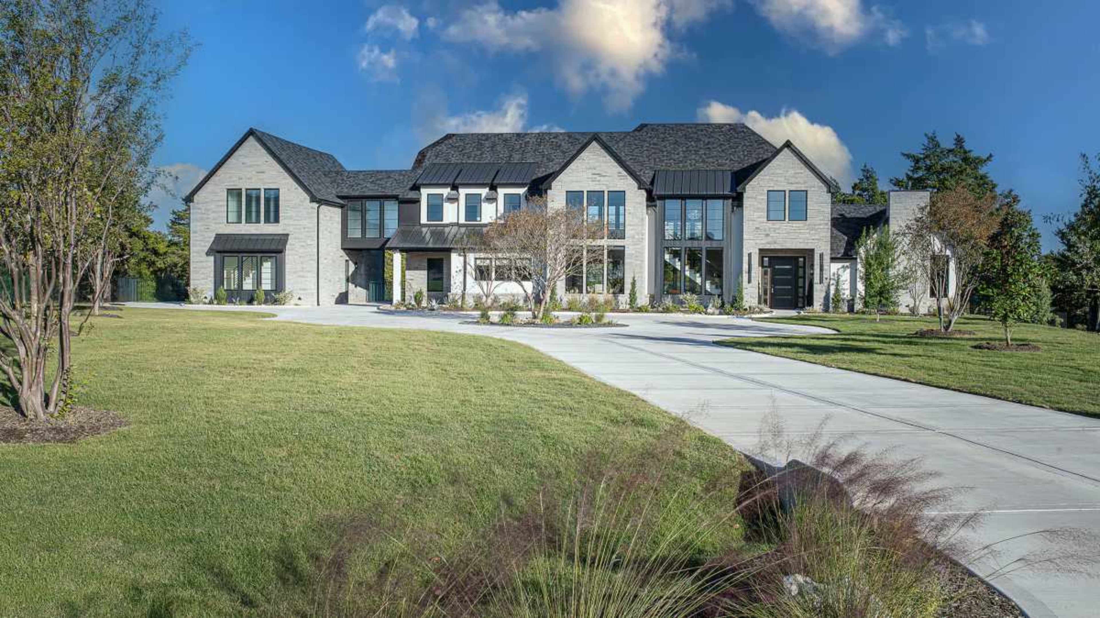 A large, modern house features a mix of stone and black metal accents, with a spacious front lawn and a concrete driveway leading to the entrance.