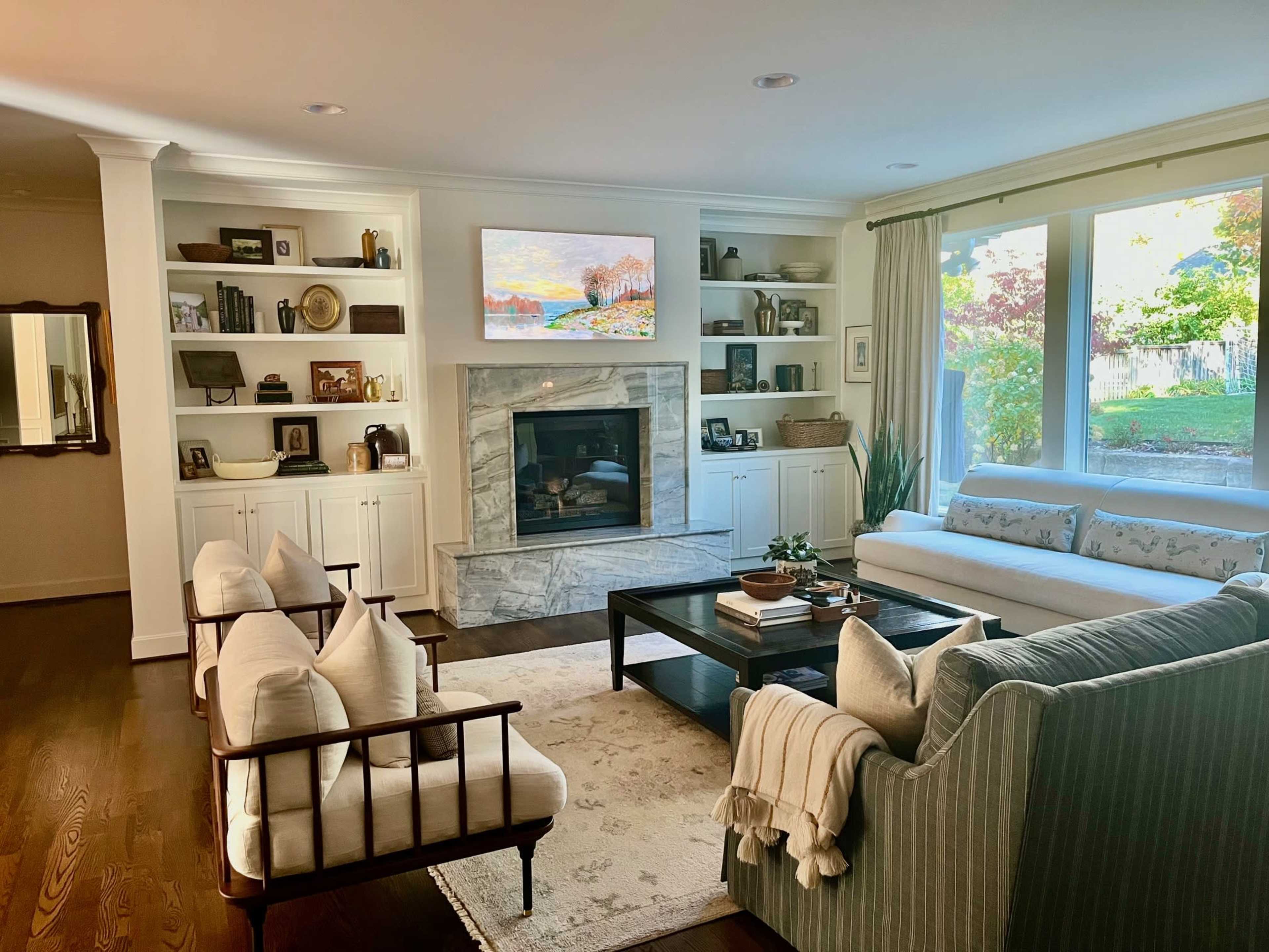 The living room features a marble fireplace surrounded by built-in shelves, a large window offering a garden view, and a combination of light-colored furniture.