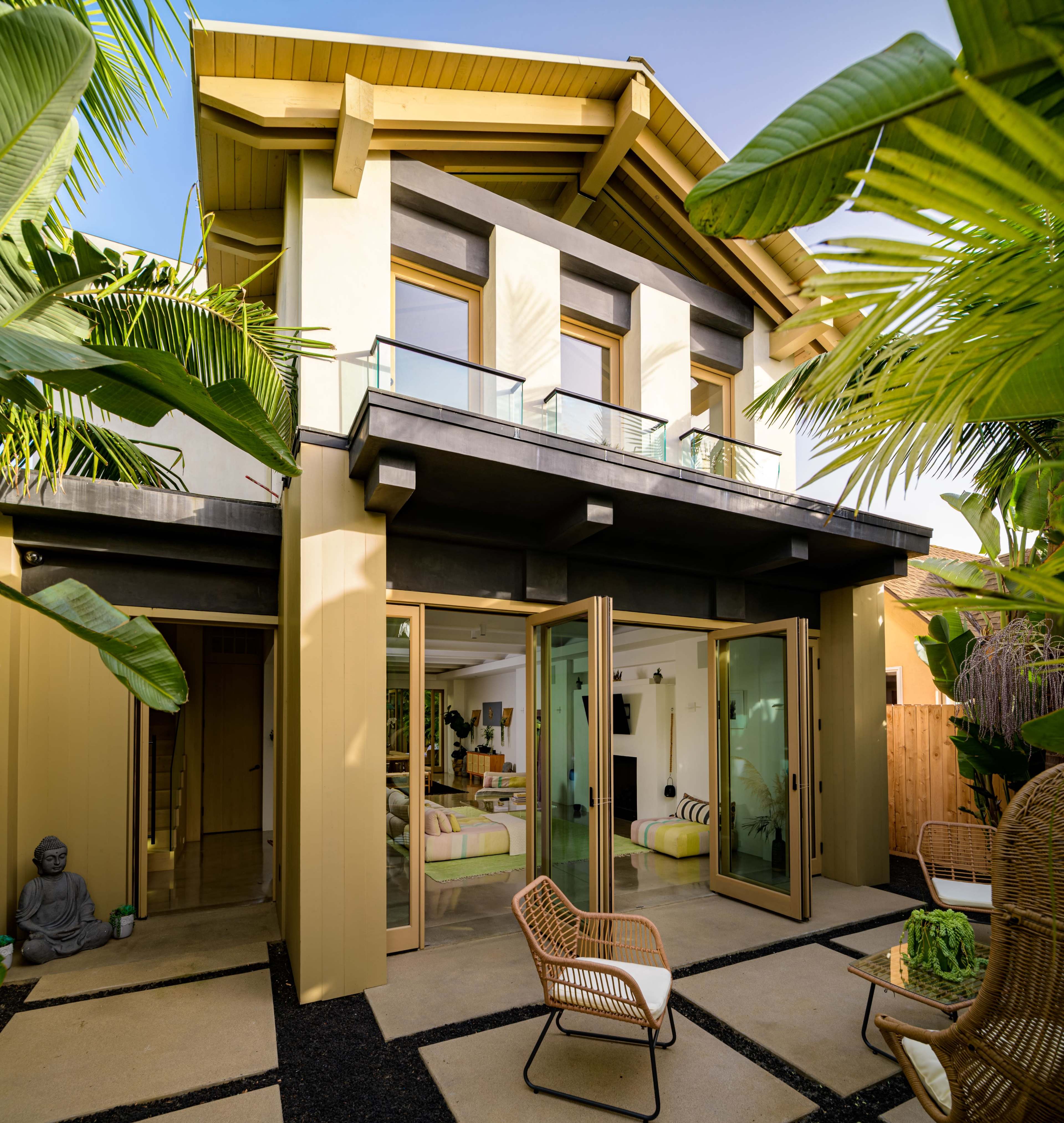 A modern two-story house with large glass doors and a balcony is surrounded by lush palm trees and a patio area.
