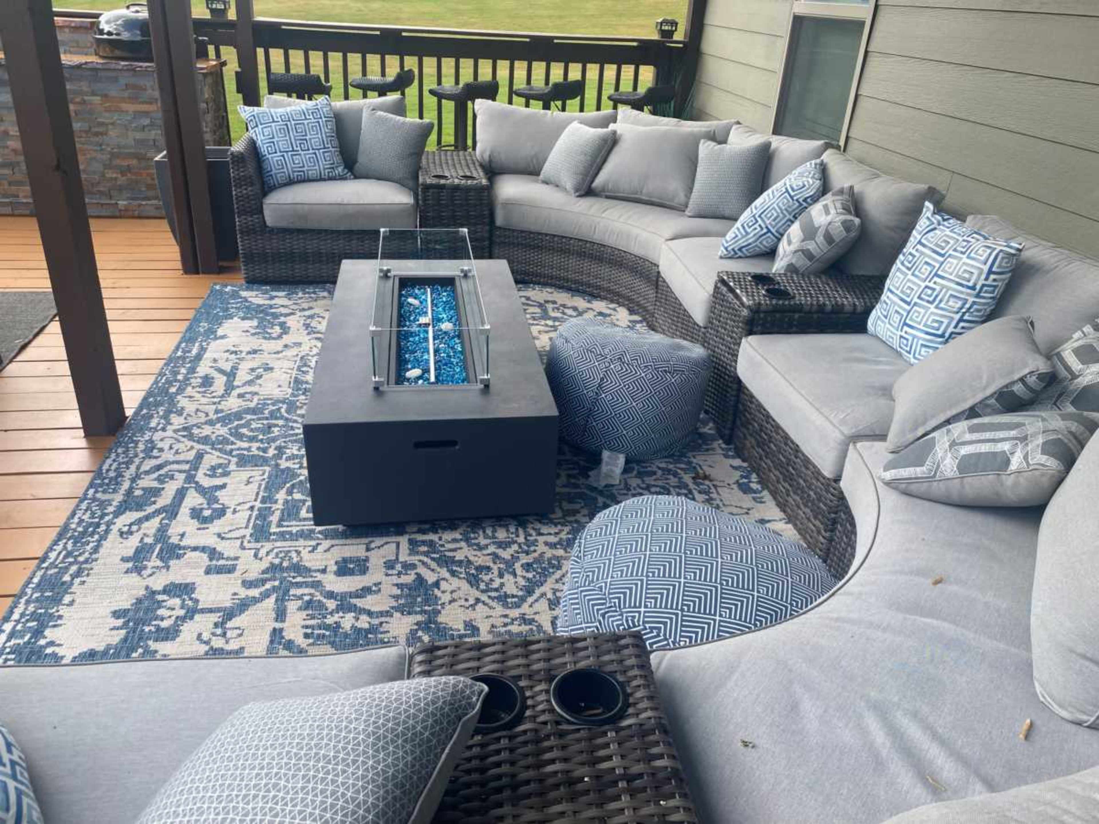 A circular outdoor seating area with gray cushions, a glass-top fire pit, and a blue-patterned rug.