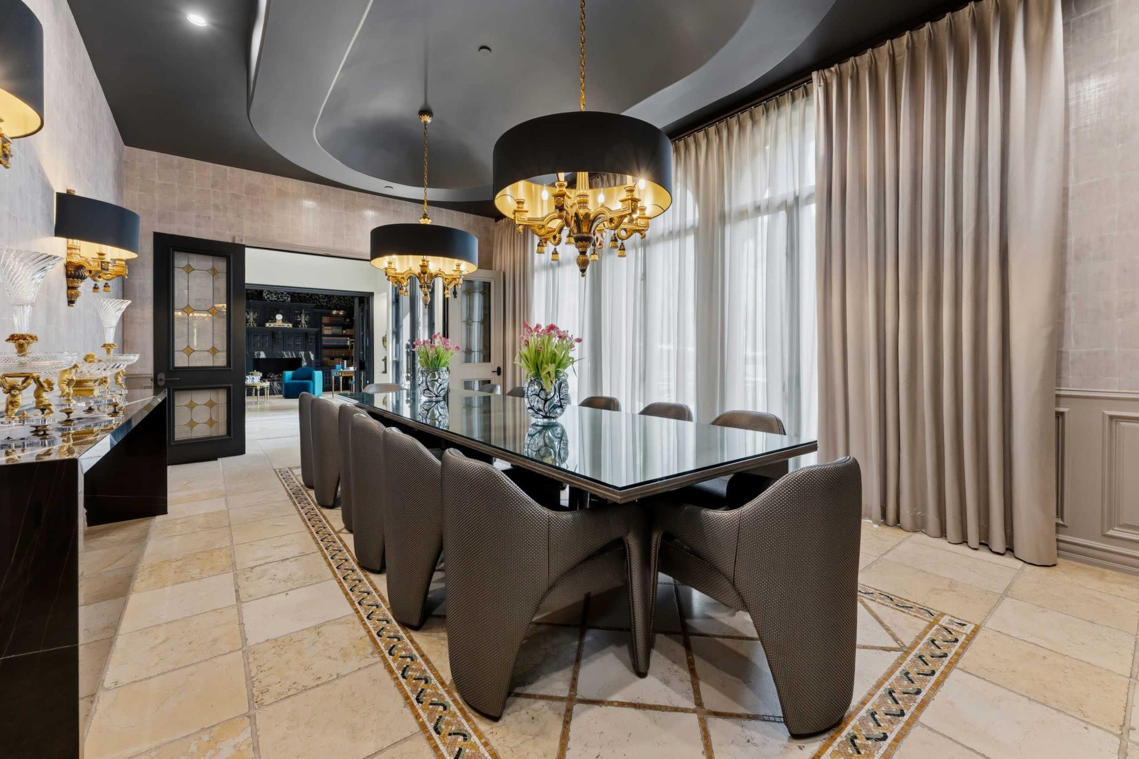 The image shows a modern dining room featuring a long glass table surrounded by upholstered chairs, with large black pendant lights and floor-to-ceiling curtains.