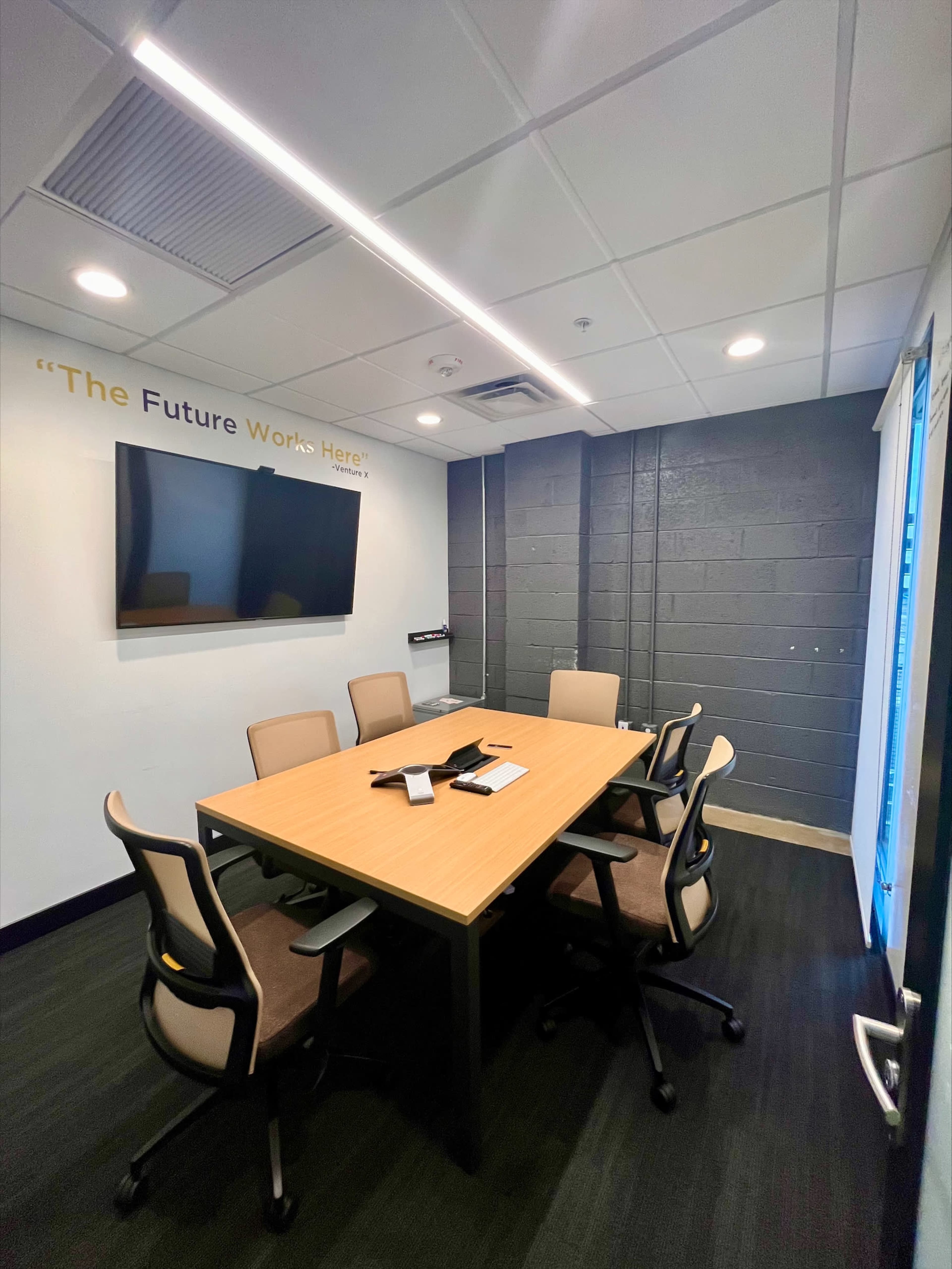 A modern conference room features a large wooden table surrounded by six chairs, a wall-mounted screen, and a slogan on the wall.