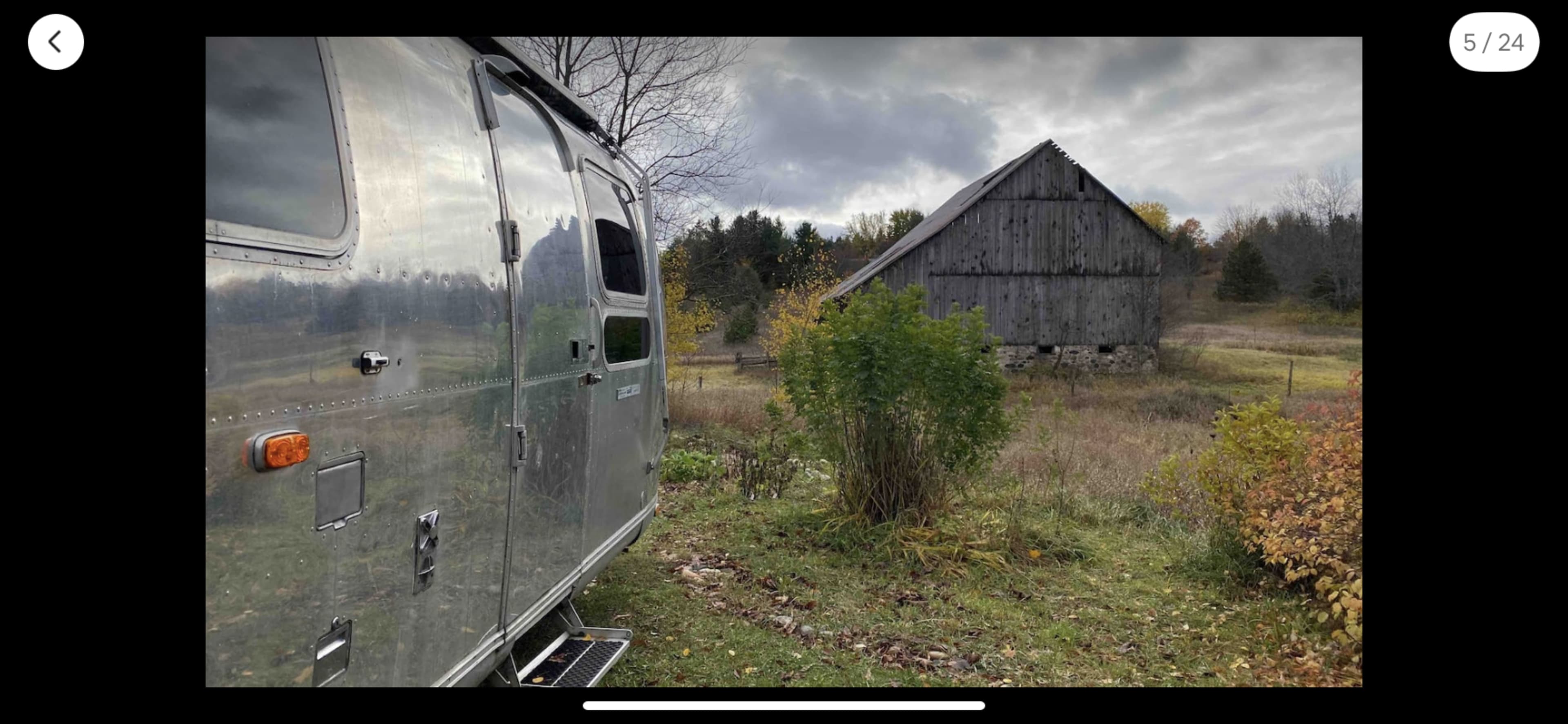 A silver Airstream trailer is parked near a weathered wooden barn in a rural landscape.