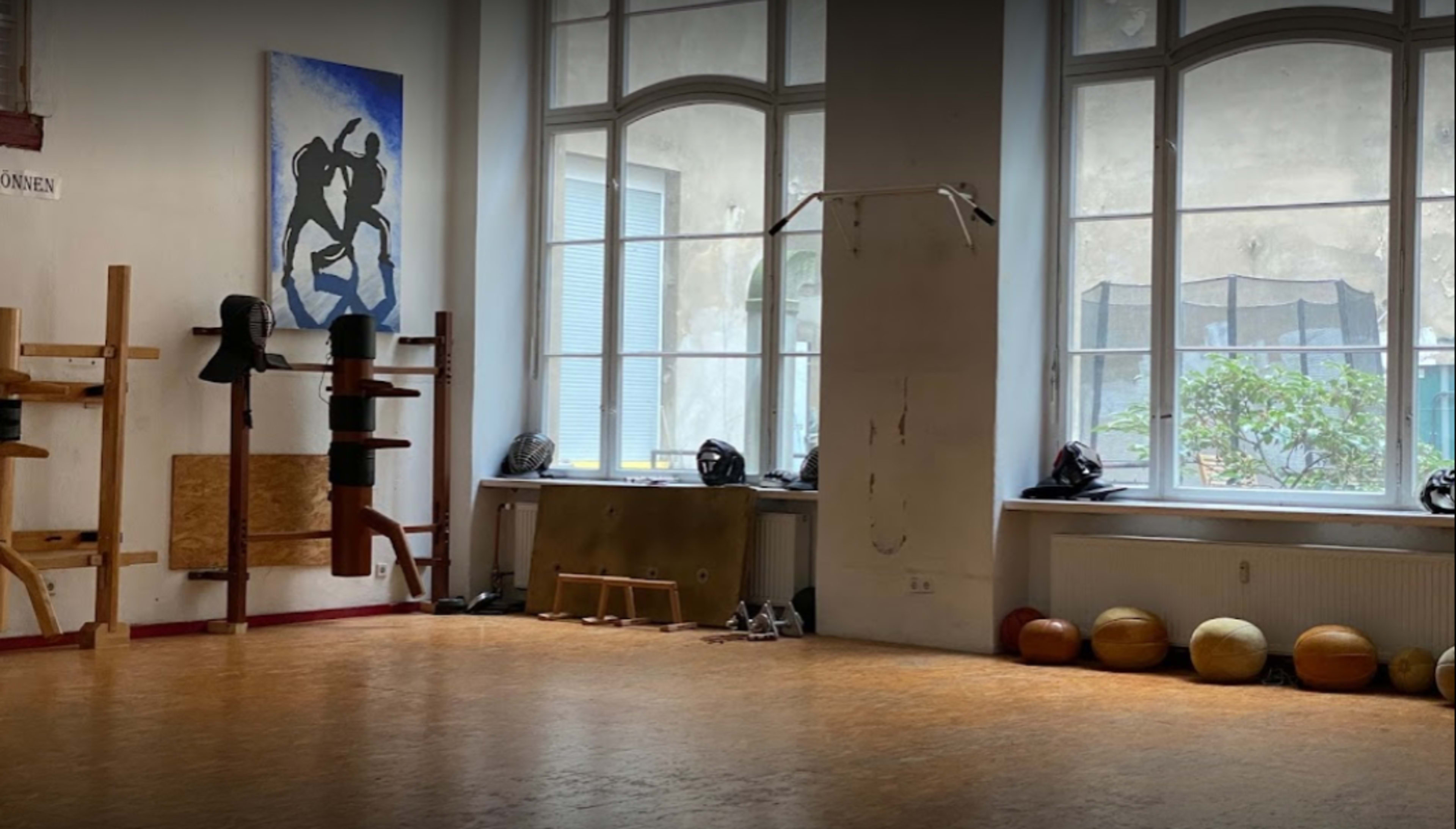 The image shows an empty training room with wooden equipment mounted on the walls, large windows allowing natural light in, and various gym balls arranged on the floor.