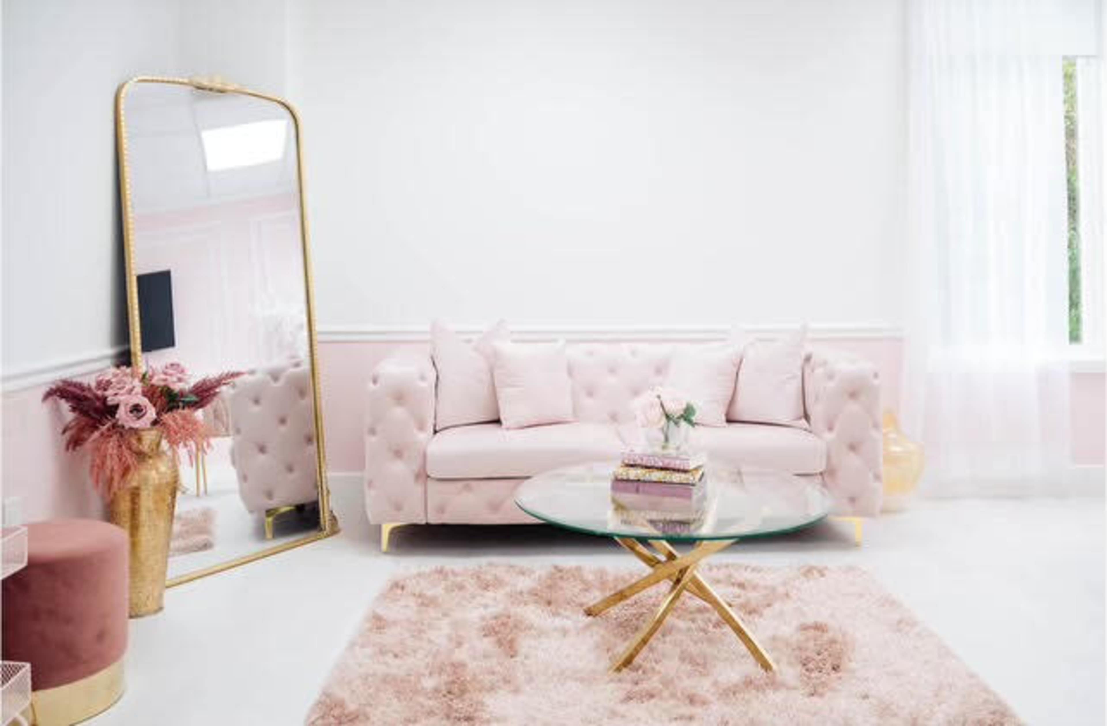 The image shows a light pink tufted sofa with pillows, a round glass coffee table, and a large mirror in a bright, softly designed living room.