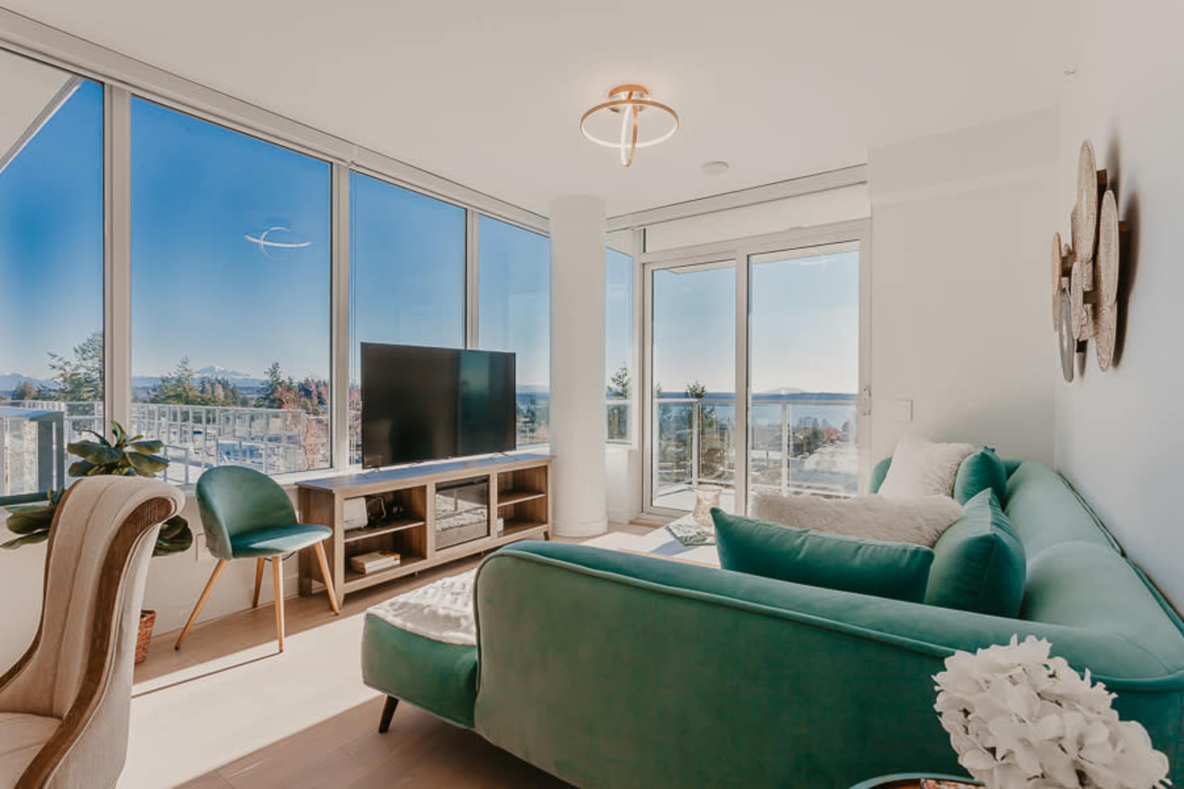A modern living room features a green sofa, a television on a wooden stand, and large windows offering a view of the outdoors.